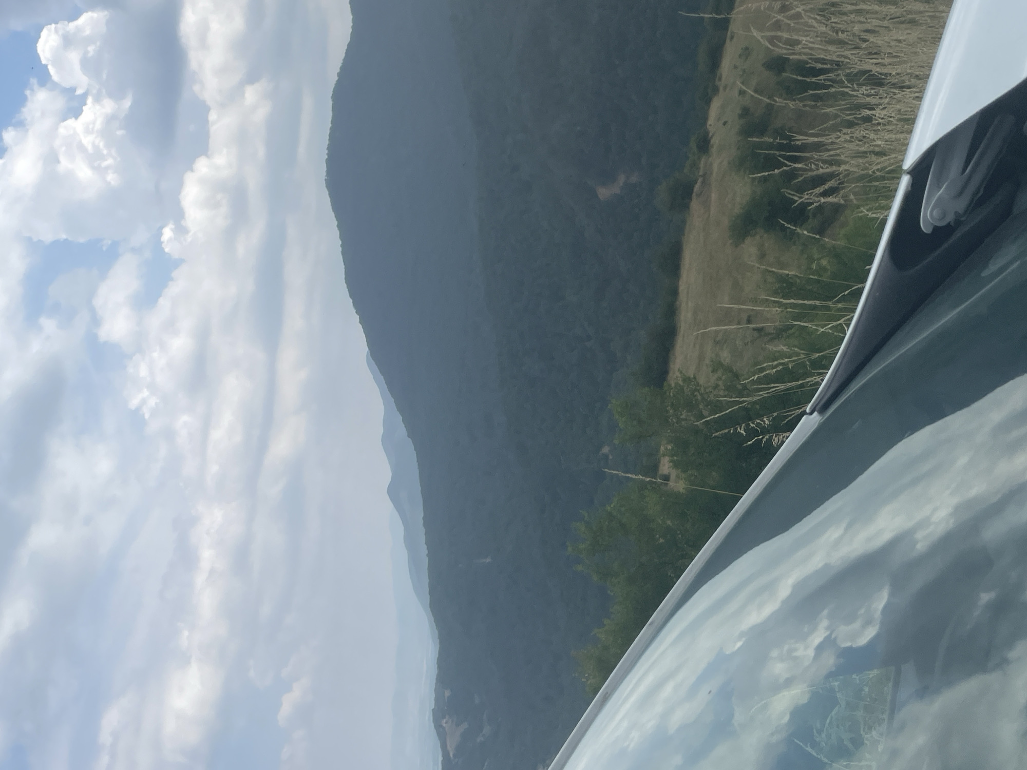 The image is a landscape photograph taken from a vehicle, looking out over a mountainous region. In the foreground, the reflection of clouds and sky is visible on the vehicle's windshield. Beyond the vehicle, rolling hills covered in dark green trees stretch into the distance, with layers of lighter, hazy mountains visible on the horizon. The sky is partly cloudy with patches of blue. The scene appears to be located in or near Oreshets, Bulgaria, given the characteristic mountainous terrain and vegetation. There are no people visible in the image.