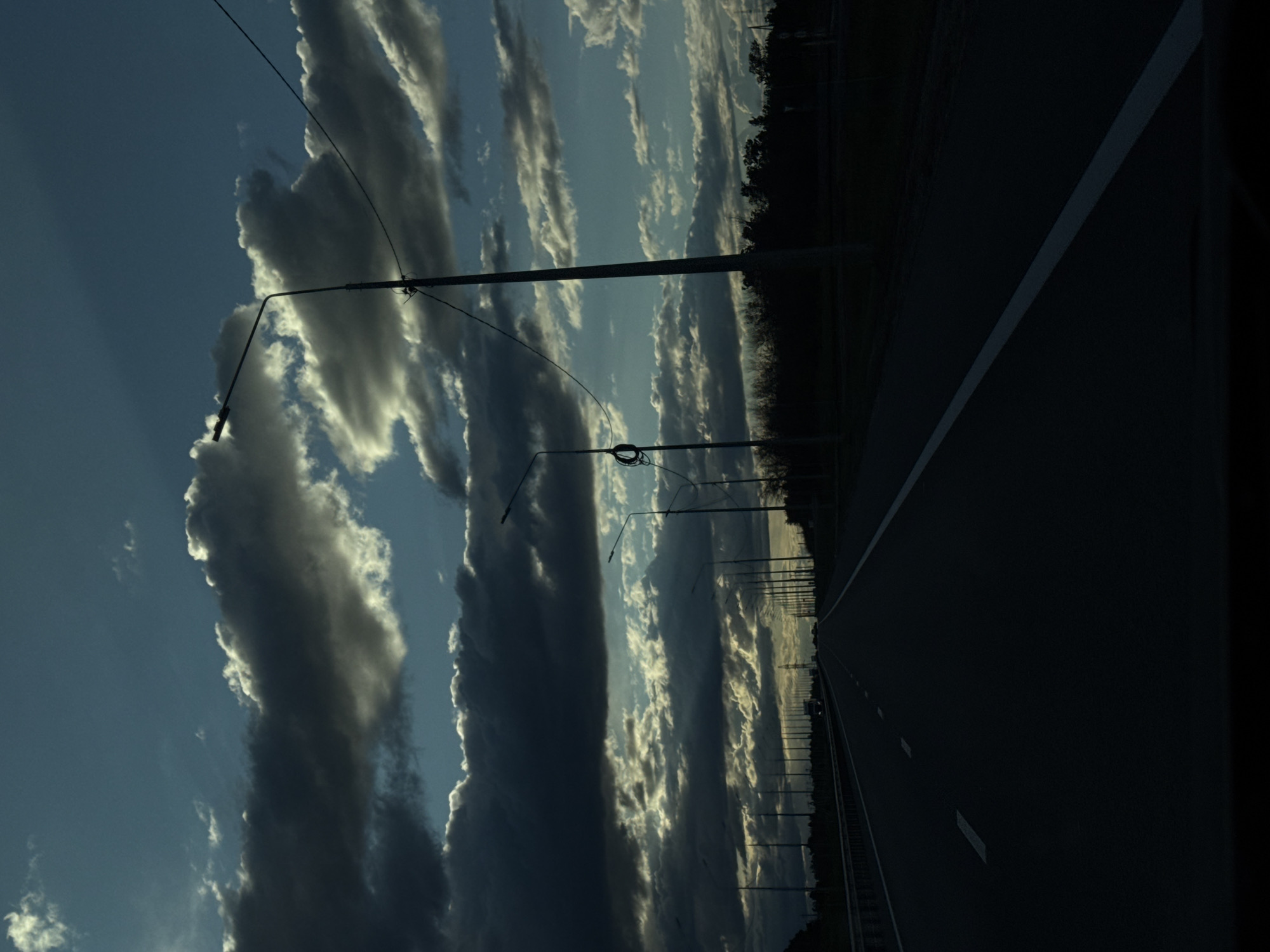The image shows a section of highway in Sokolovyi, Russia. The right side of the frame shows the dark asphalt road with a white dividing line. A dark row of trees and shrubs runs along the side of the road. On the left, the sky is visible with rows of elongated dark and white clouds. Streetlights stand alongside the road, extending into the sky.