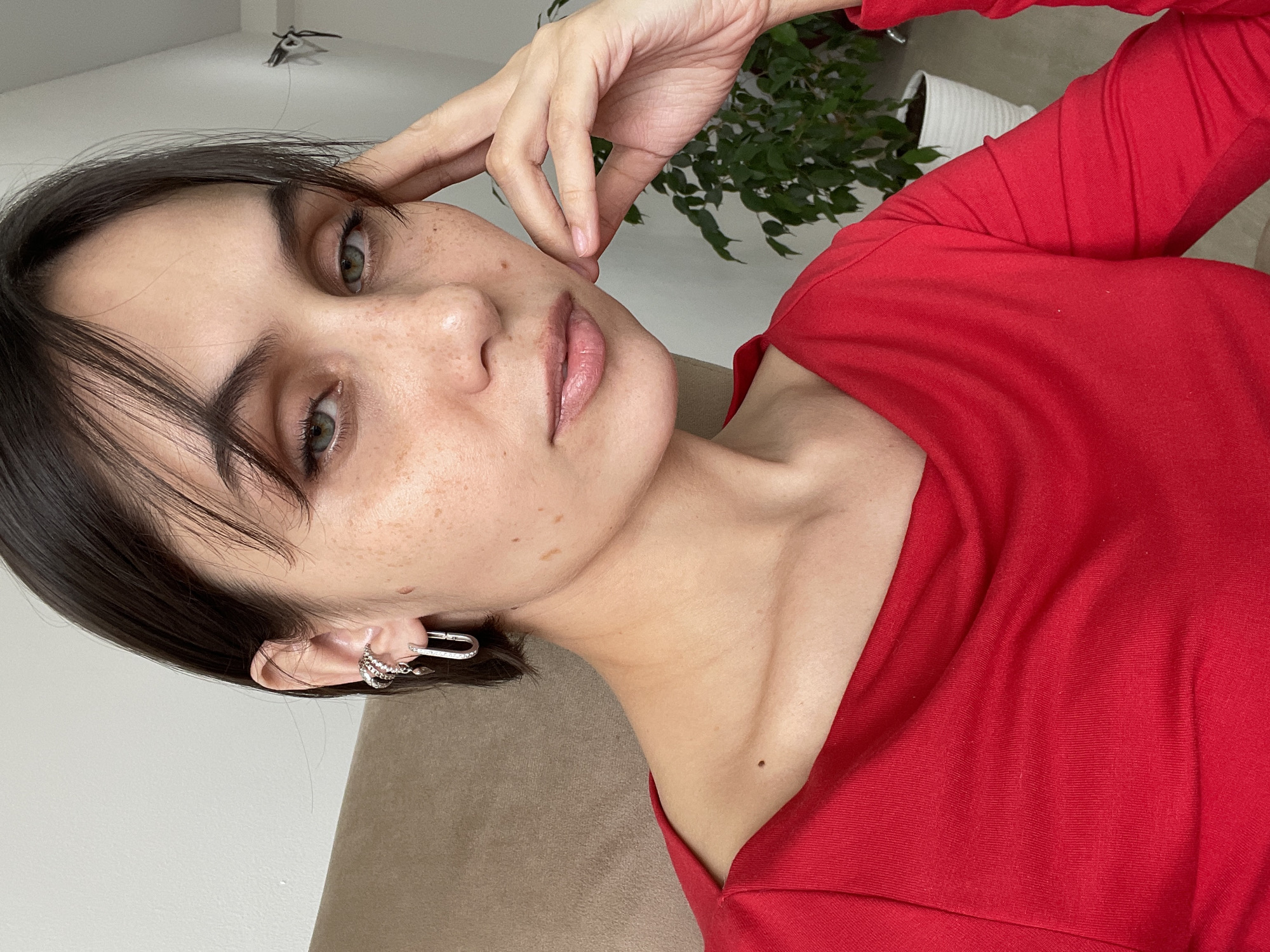 A woman with dark hair and fair skin, freckled, is wearing a red long-sleeved shirt. She has green eyes and is wearing silver earrings. Her hand is near her face. A green plant is visible in the background. The location is inside a building. It is not possible to determine the specific location in Izhevsk, Russia from this image.