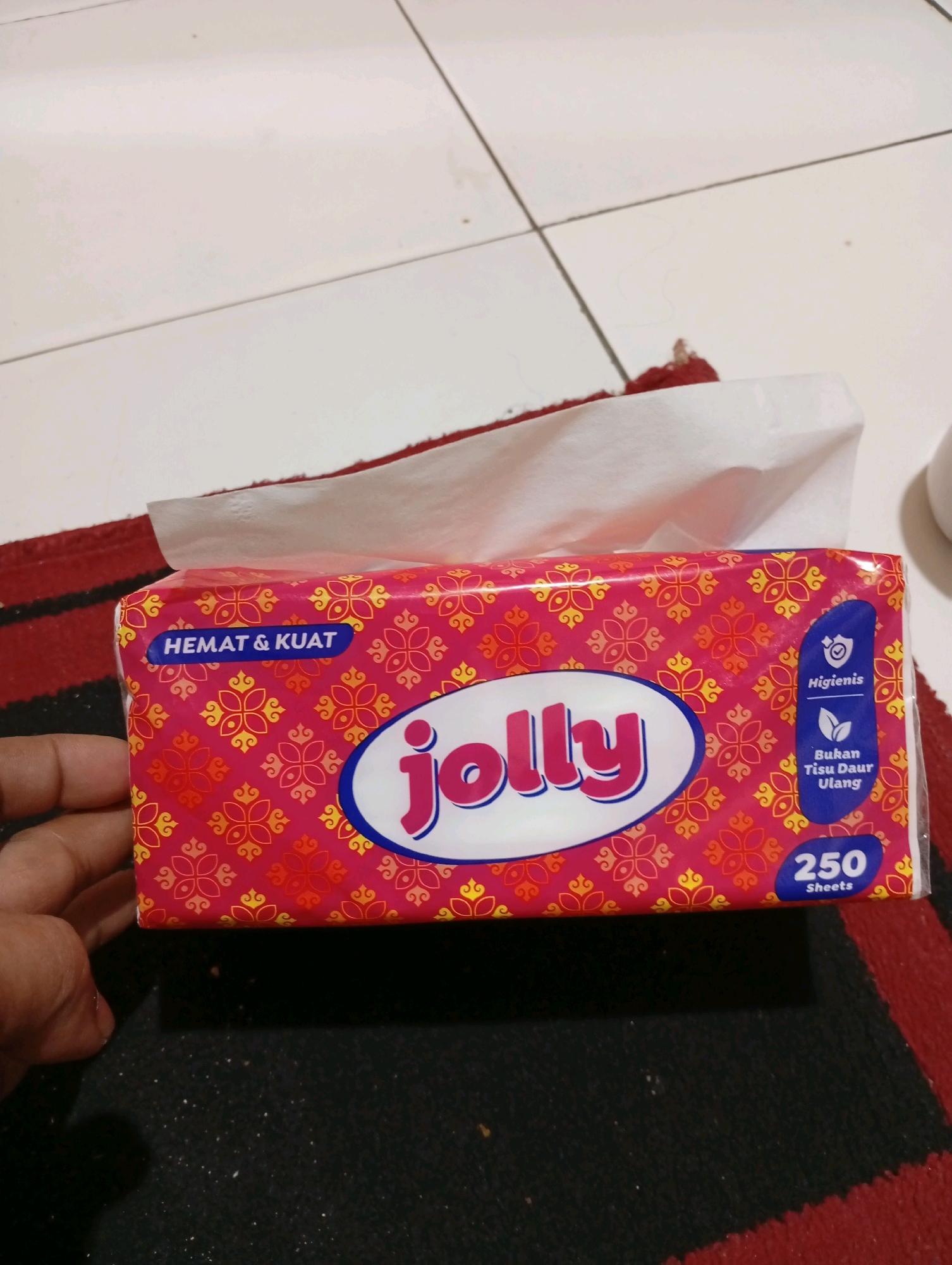 A frontal, eye-level, medium shot captures a pink box of "Jolly" brand facial tissues placed on a dark red and black striped rug. The box features a repeating floral pattern in gold and orange. Text on the box includes "HEMAT & KUAT" in blue on the left, the "Jolly" logo in an oval in the center, and "Higienis" with an icon of a shield with a checkmark, and "Bukan Tisu Daur Ulang" (Not Recycled Tissue) with a leaf icon on the right. Below this, the number "250 Sheets" is displayed in a blue oval. The top edge of the box is slightly open, revealing white tissue. A person's hand, partially visible on the left side of the frame, is holding the box. The background consists of a white tiled floor. The scene is indoors, likely in a residential setting. It is not possible to determine the specific location within the city of Caringin, Indonesia from the image alone.