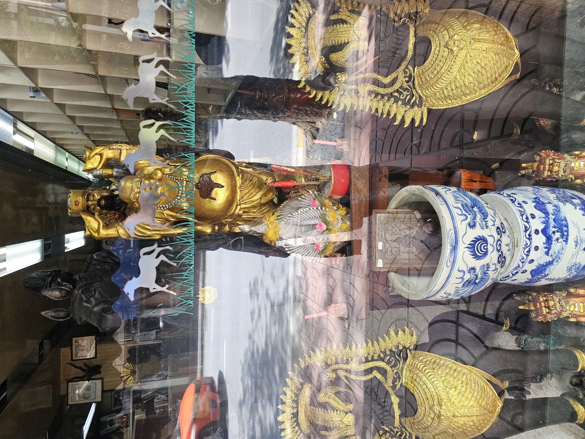This image captures a street-level view through a shop window in Bangkok, Thailand, offering a glimpse into a space filled with religious and decorative artifacts. The foreground is dominated by large, dark statues adorned with elaborate gold headdresses and breastplates, possibly representing mythical beings or deities. Between these statues, a tall, golden statue of a figure in traditional attire stands prominently, holding various symbolic objects and wearing a heavy beaded necklace.
In front of the golden statue, a red pot holds incense sticks, with a floral offering beside it. Further forward, a two-tiered ceramic pot, decorated with blue and white dragon motifs, sits on a wooden surface. A small, framed document with intricate diagrams and text is placed to the left of the ceramic pot.
The window reflects the street scene outside, including the blurred image of a red car driving past, a tree trunk, and pavement tiles. The background within the shop appears to be shelves filled with various items, though they are obscured by reflections and the objects in the foreground. The overall impression is one of a vibrant, culturally rich environment, possibly a shop specializing in religious items, amulets, or traditional crafts, situated on a busy Bangkok street.