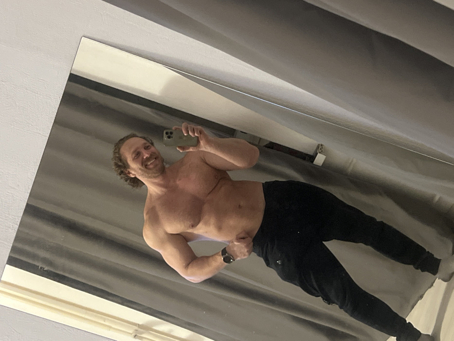 A man with curly brown hair smiles, holding a phone in his right hand to take a mirror selfie. He is shirtless, revealing a muscular physique. He wears black sweatpants and a watch on his left wrist. The background consists of a gray curtain and part of a white ceiling, which are seen in the mirror. The image is angled as if the mirror itself is tilted.