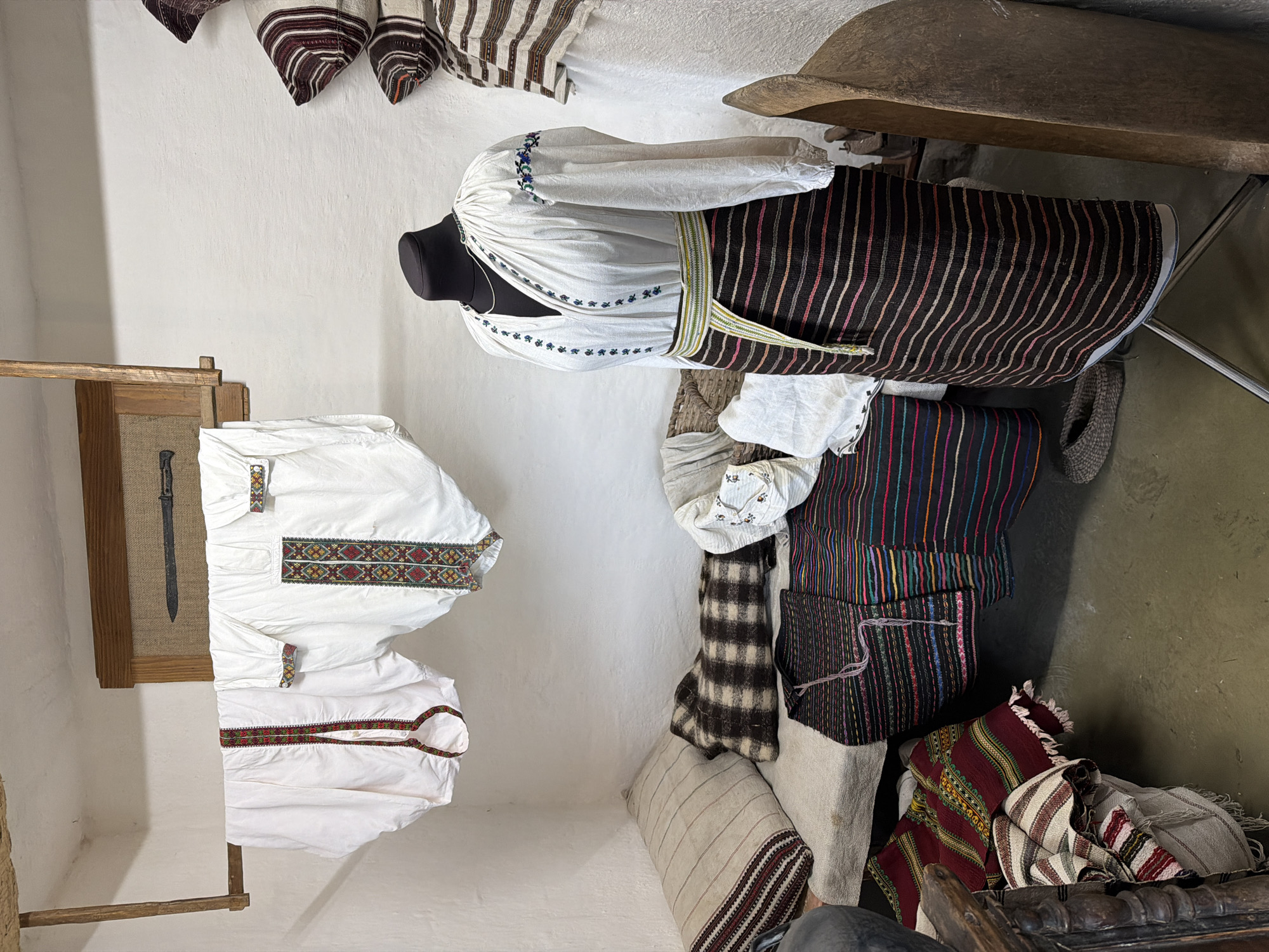 This image depicts an indoor scene, likely a museum exhibit or a curated display of traditional clothing, located in Velykyi Khodachkiv, Ukraine. The room has white walls and a light-colored floor. In the foreground, a mannequin is dressed in a white embroidered blouse and a striped skirt, cinched with a woven belt. The blouse features intricate embroidery around the neckline and cuffs.
Behind the mannequin, several pieces of white fabric, possibly shirts or undergarments, are hung on a wooden rod suspended from the ceiling. These garments are also adorned with colorful embroidered trim. To the left of the mannequin, a rustic bed or bench is covered with striped pillows and blankets. Folded textiles with vibrant striped patterns are piled on a wooden structure in the lower left corner. To the right of the mannequin, a large, curved wooden object, possibly a trough or a boat-shaped vessel, stands upright.
On the wall behind the hanging garments, a framed display contains what appears to be a historical weapon, such as a sword or a dagger. The overall impression is one of a preserved collection showcasing traditional Ukrainian folk attire and domestic artifacts.