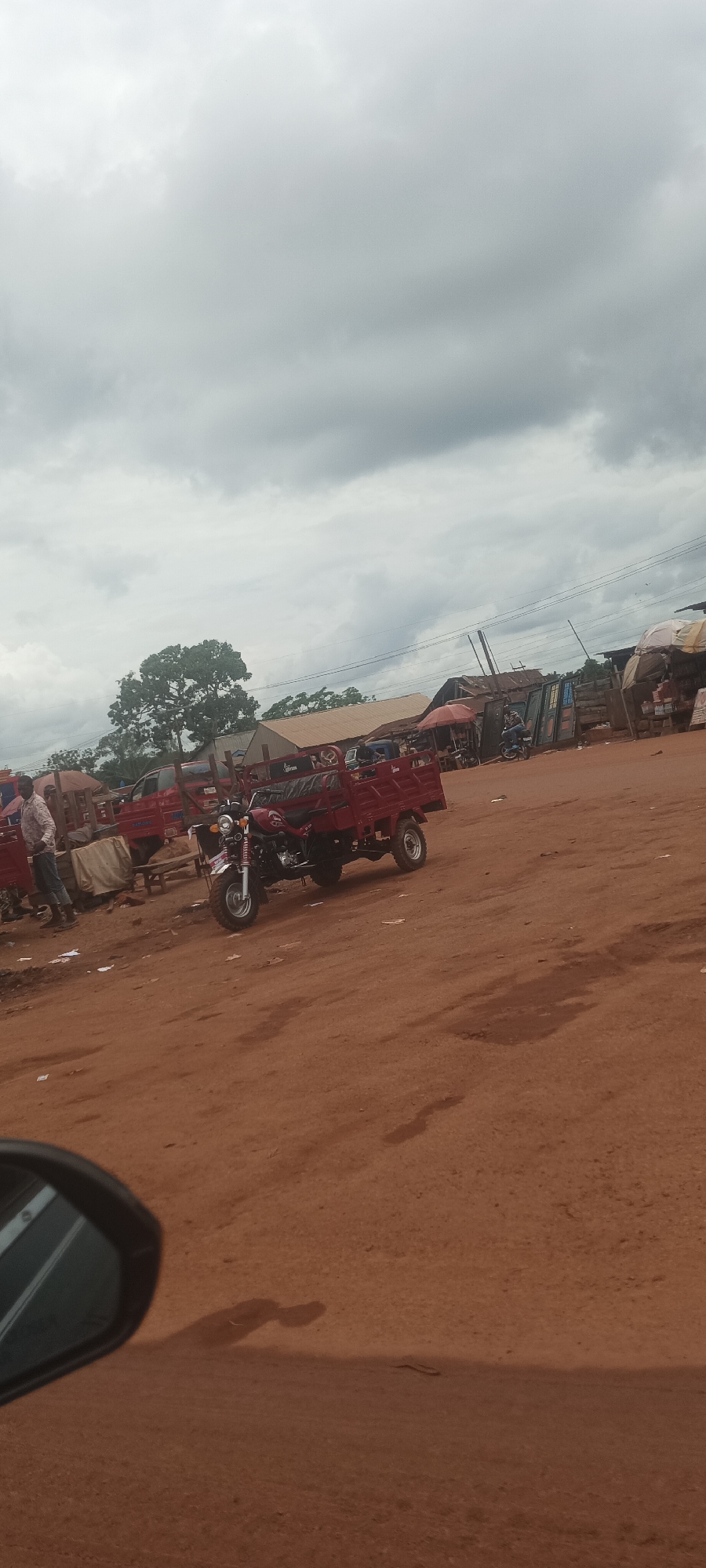 The image captures a scene in Imilike, Nigeria, characterized by a reddish-brown dirt ground and overcast sky. In the foreground, a three-wheeled motorcycle/truck hybrid vehicle is parked. Other similar vehicles and structures which appear to be small shops or stalls are visible in the mid-ground. A man is seen standing near one of these vehicles. In the lower left corner, a car mirror is partially visible. The environment is dusty, open, and suggests a market or transportation hub.