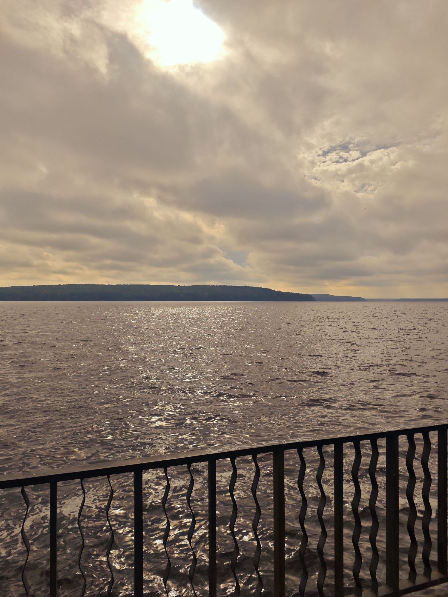 The image shows a body of water, likely the Kama River in Chaikovsky, Russia, as viewed from a promenade. A dark metal railing with a decorative wavy pattern runs across the foreground, obscuring the lower portion of the water. The water's surface reflects the sunlight, creating a shimmering effect. In the distance, a dark, tree-covered shoreline stretches across the horizon. The sky above is overcast with large, textured clouds, partially obscuring a bright, sunlit area. The overall atmosphere is muted, with warm, diffused light.