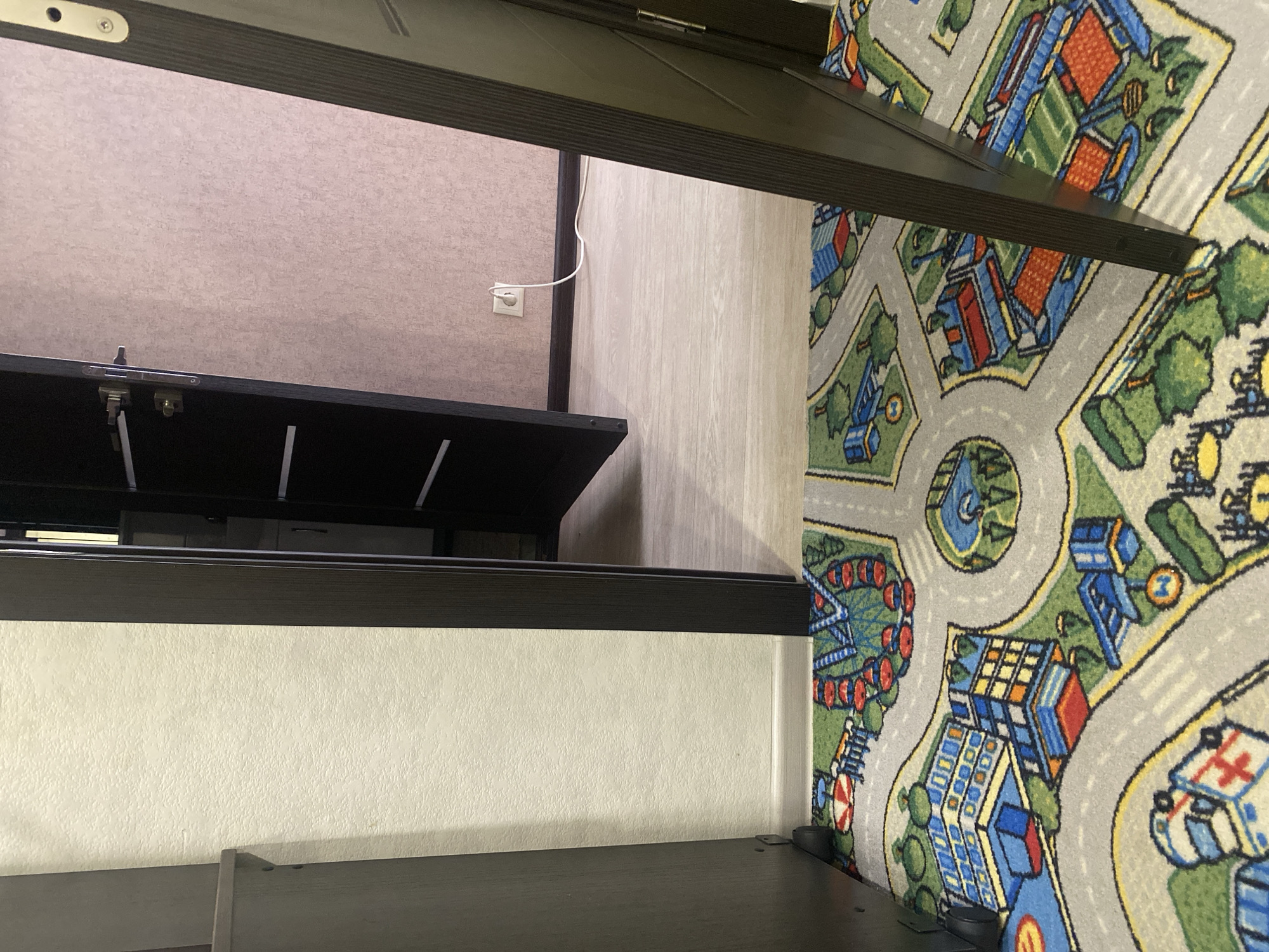 The image shows an interior hallway with a dark brown door ajar, revealing another room beyond. A light-colored wall is visible on the left, and the floor is covered by a colorful children's rug depicting a city street scene with buildings, cars, and a Ferris wheel. The rug has a grey base with intricate patterns in green, blue, red, and yellow. In the background, a light wooden floor can be seen. A white electrical cord runs from a wall outlet on a light pink textured wall to an unseen appliance. The scene is well-lit, suggesting daytime. There are no people present in the image. The location is not specified to be in Saratov, Russia or any other city.