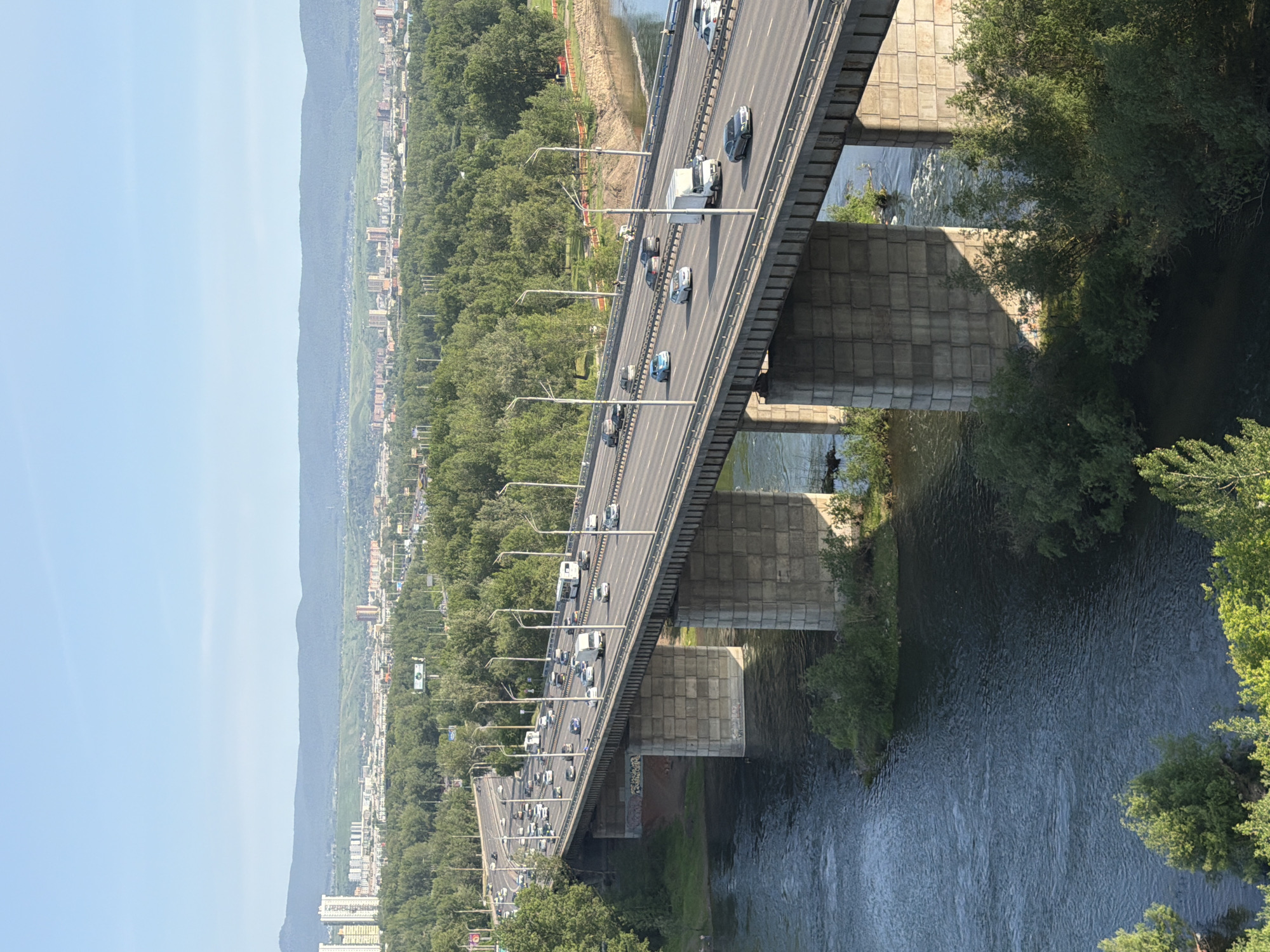 The image displays a wide, multi-lane bridge spanning a dark, flowing river. The bridge, constructed with concrete pillars and supports, carries a significant volume of traffic, including cars and a white truck. Trees and lush green foliage line the riverbanks and the landscape beyond the bridge, which recedes into a hazy, mountainous terrain dotted with buildings, suggesting an urban or suburban environment. The sky is a clear, pale blue, indicating a bright, sunny day. The perspective is from a high vantage point, looking down and across the bridge, emphasizing its scale and the surrounding natural and man-made elements. The scene is serene and busy, with the river adding a natural element to the infrastructure of the bridge and the distant city. No visible text is present in the image.