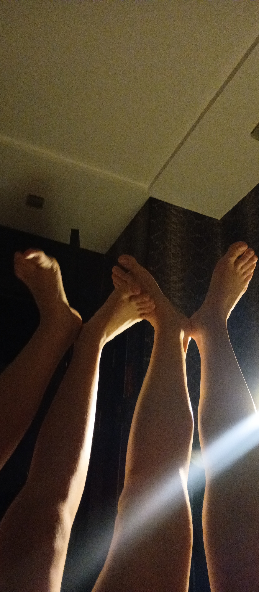 The image depicts the lower half of three individuals, specifically their legs and feet, positioned upwards against a ceiling and wall. The ceiling is a light color, possibly off-white, with a recessed light fixture visible. The wall is covered in a textured dark material. The legs are of varying shapes and sizes and appear to be fair-skinned. A bright light source illuminates the legs on the right side of the frame. The location is indicated as Unknown, Unknown.