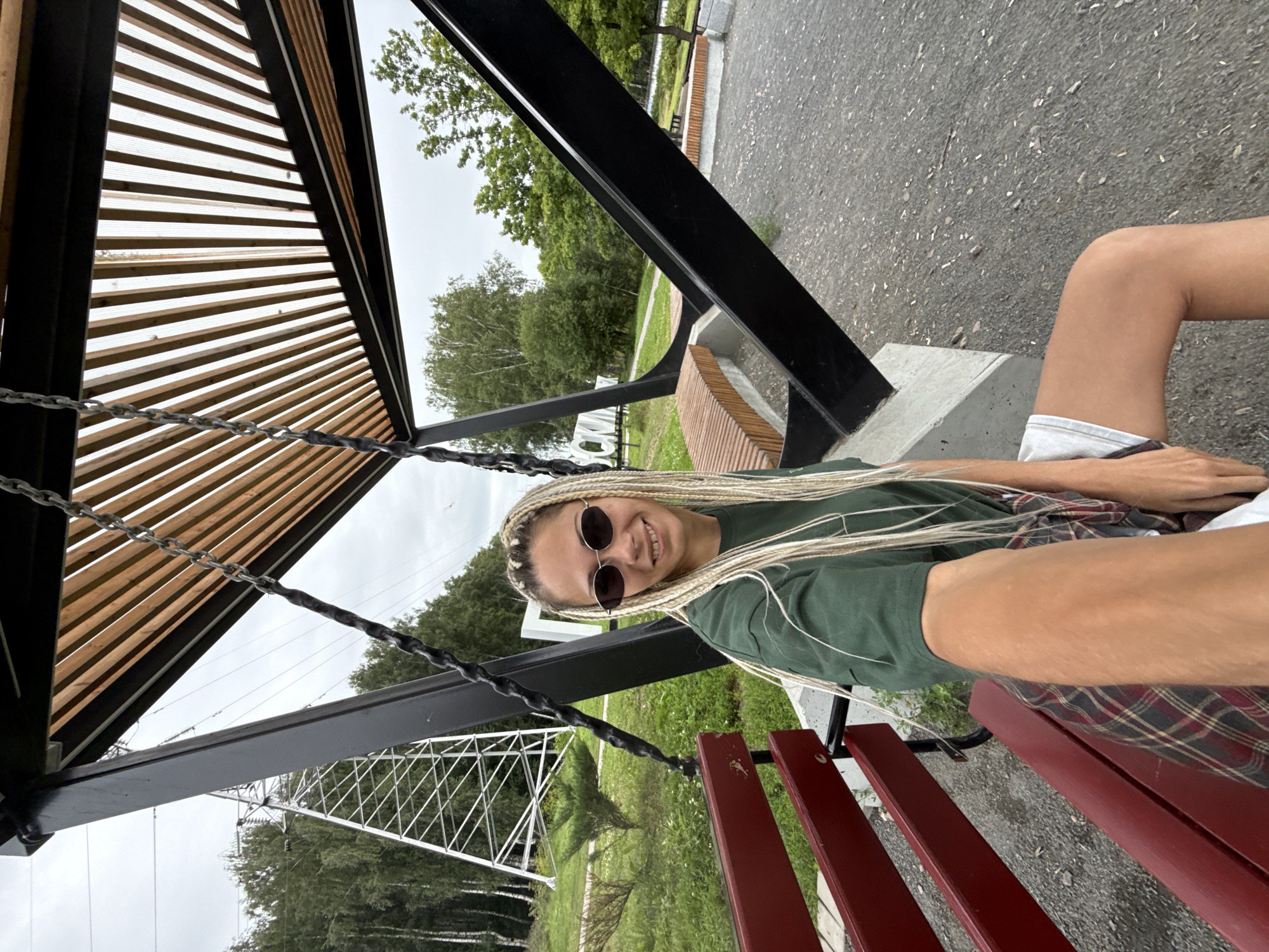 In the city of Rusanovo, Russia, a young woman with blonde braided hair and sunglasses is sitting on a red bench under a modern outdoor structure with a wooden slatted roof. She is wearing a green t-shirt, a plaid shirt tied around her waist, and light-colored shorts. The structure appears to be a modern swing set or seating area. In the background, a large white power pylon and a forest of trees are visible. The ground is covered with gravel.