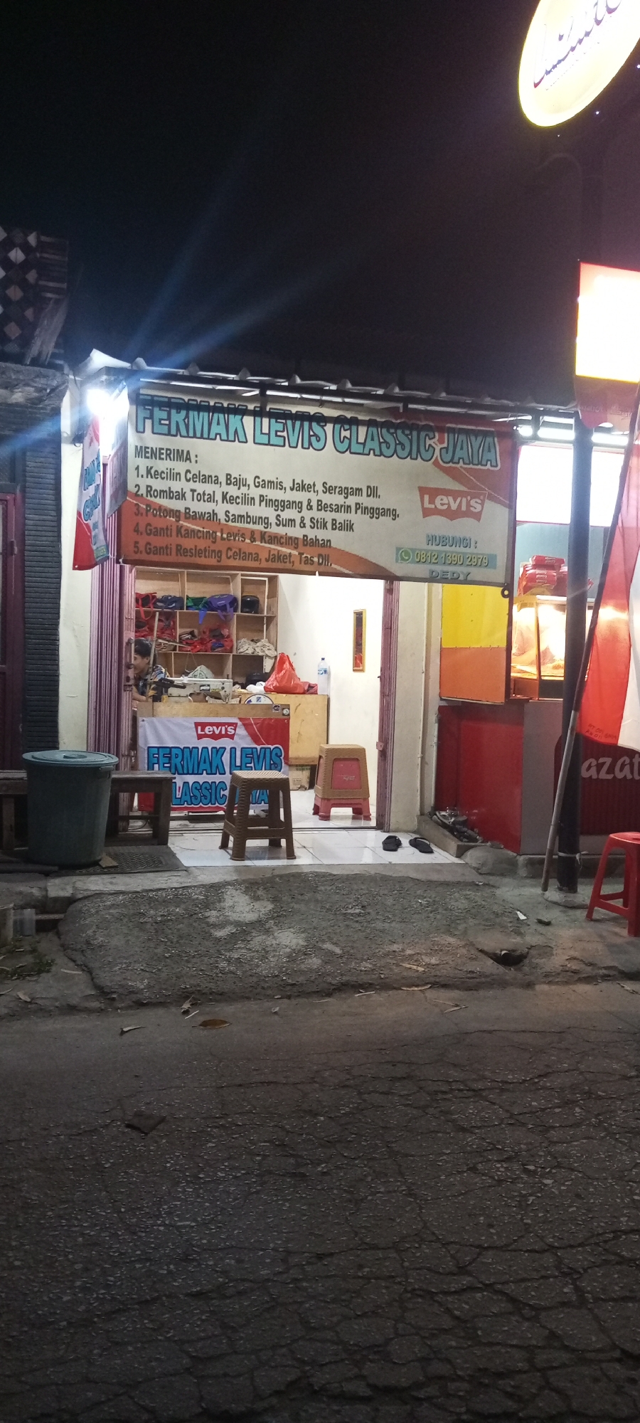 The image captures a nighttime street view of a shop named "FERMAK LEVIS CLASSIC JAYA" located in Setu, Indonesia. The shop's facade is illuminated by artificial lights, with a prominent sign bearing the shop's name and a Levi's logo. Below the main sign, a list of services is displayed, including "Menerima: 1. Kecilin Celana, Baju, Gamis, Jaket, Seragam Dll. 2. Rombak Total, Kecilin Pinggang & Besarain Pinggang. 3. Potong Bawah, Sambung, Sum & Stik Balik. 4. Ganti Kancing Levis & Kancing Bahan. 5. Ganti Resleting Celana, Jaket, Tas Dll." A phone number, "0812 1390 2979", is also visible, associated with the name "DEDY".
Inside the shop, shelves filled with various items, possibly clothing materials and accessories, are visible. A person, appearing to be a young male, is seated inside the shop, seemingly occupied with work. In front of the shop, there are several objects, including a large green trash bin, wooden benches, and plastic stools. The street in front of the shop is paved with asphalt and shows signs of wear and tear, with cracks and debris scattered on the surface. A red stool and a black pole with Indonesian flags are also present near the shop's entrance. The overall scene depicts a typical street-level commercial establishment operating at night in an Indonesian urban environment.