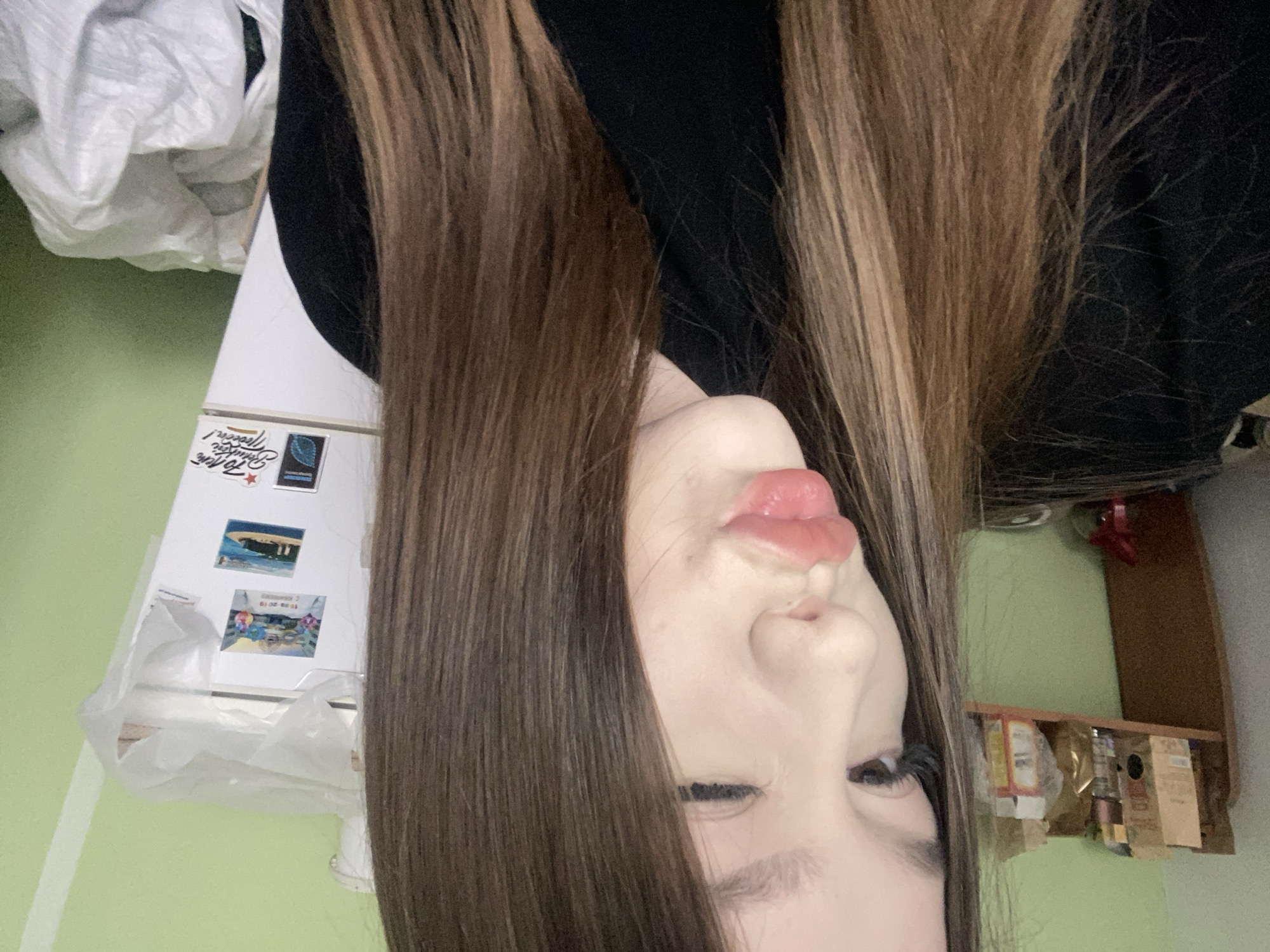 A selfie image shows a person with light skin, brown hair with blonde highlights, wearing a black garment. The person's face is positioned in the center of the frame, with their lips pursed slightly. The background includes a refrigerator with magnets and a white plastic bag, a light green wall, and a brown shelf with various items. The location is likely Chaikovsky, Russia, based on the refrigerator magnets.