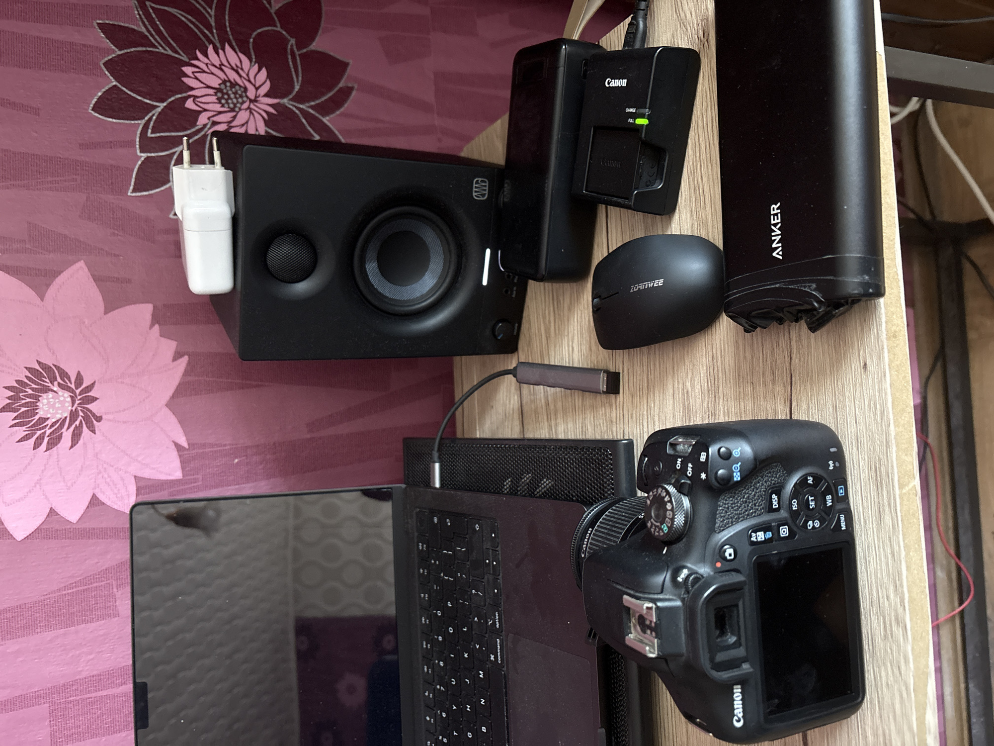 The image shows a cluttered desk surface with various electronic devices. A black speaker with a white AC adapter plugged into the side is on the left. Next to it are a Canon battery charger, a black power bank labeled "ANKER", and a small black computer mouse labeled "ZOROWEE". A laptop with a black keyboard is in the lower left. A Canon DSLR camera with a lens attached is on the lower right. The background includes a floral patterned wall and a light-colored wooden surface. The location in the city of Lukovit, Bulgaria is not discernable from the image.