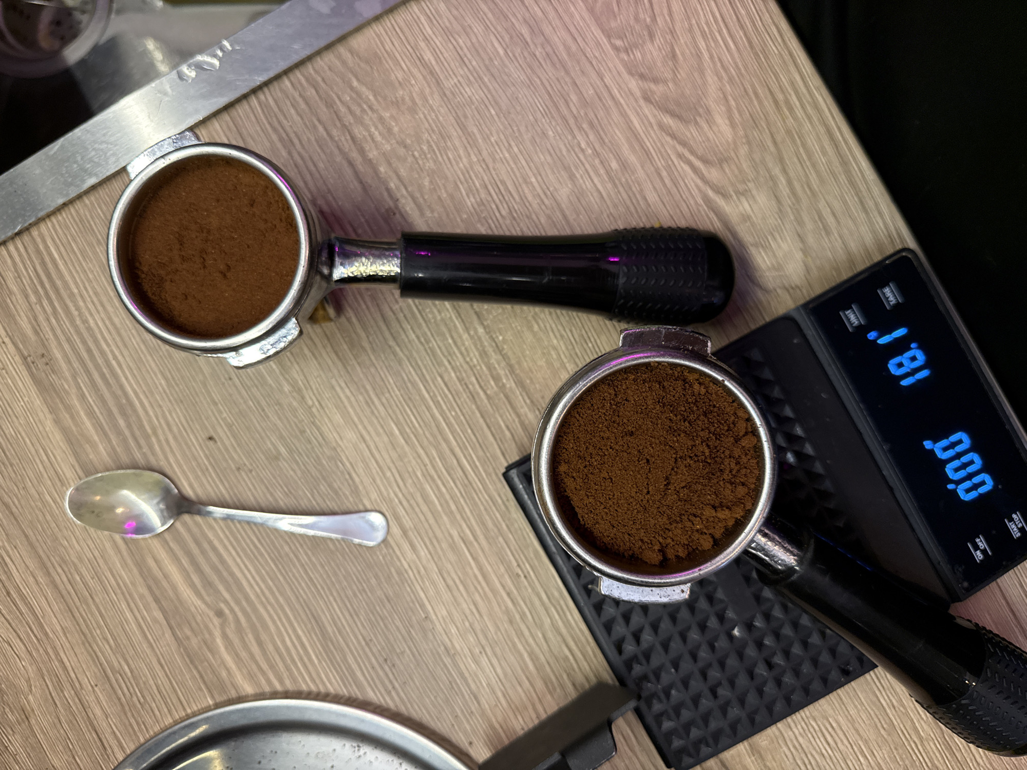 An overhead, close-up shot captures a coffee preparation scene on a light brown, wood-grain countertop. In the upper portion of the frame, a silver spoon rests on the counter. Below the spoon, a portafilter filled with finely ground coffee is positioned to the right. Further down and to the left, another portafilter, also filled with coffee grounds, sits on a black, textured scale. The scale's digital display reads "0.00" and "18.1°". A portion of a silver, rectangular object is visible on the right side of the image, potentially a part of a coffee machine or a tamper. The scene is illuminated with subtle pink and purple lighting, creating reflections on the metallic surfaces.