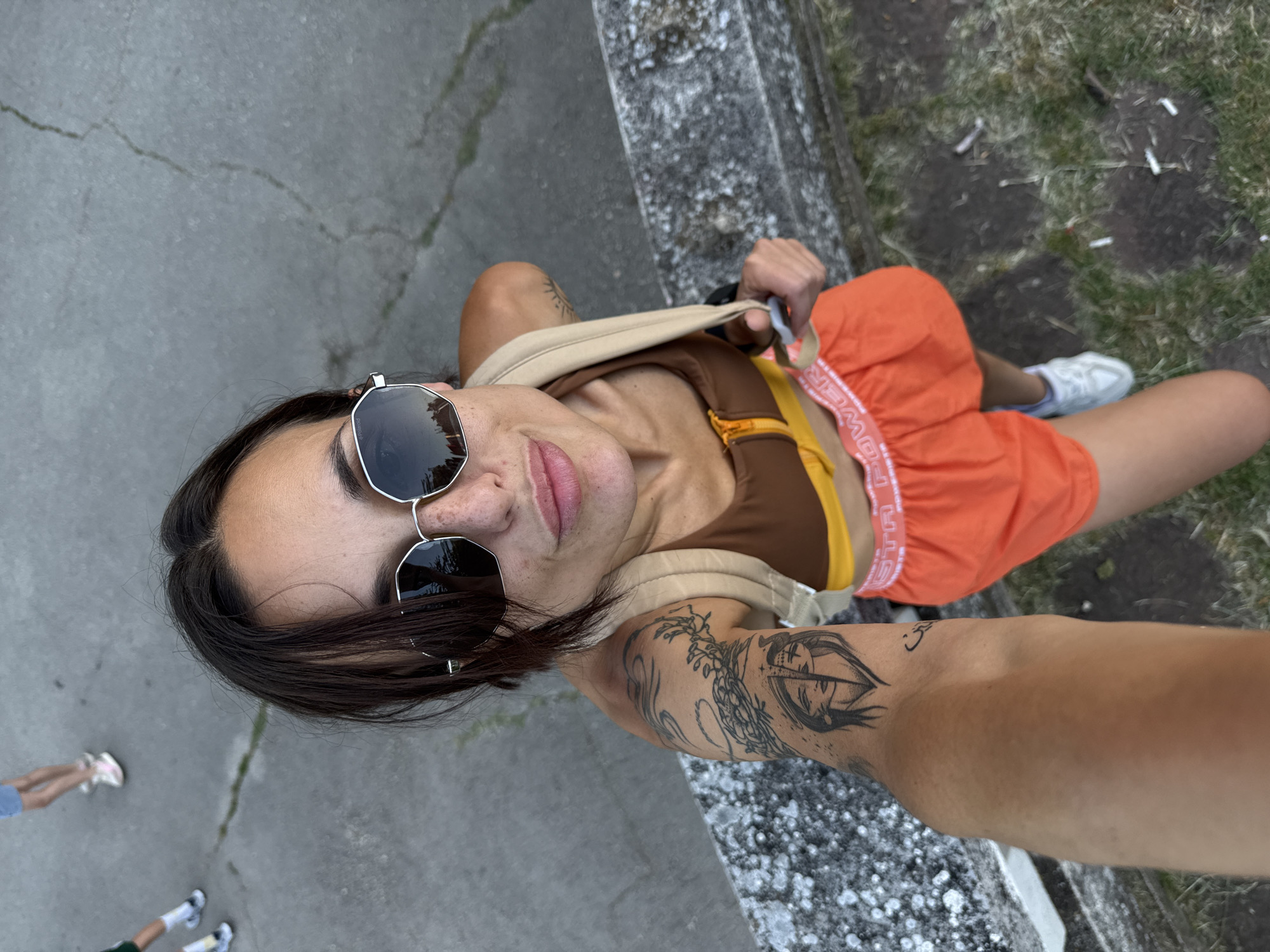 The image shows a high-angle selfie of a young woman wearing sunglasses, a brown and yellow athletic top, and orange shorts with the word "POWER" visible on the waistband. She has a tattoo on her left arm. She is standing on a paved surface next to a stone ledge, with grass and cigarette butts on the ground below. In the background, out of focus, are the legs of two other individuals. The scene appears to be outdoors during daylight. The overall context does not provide any specific indication of the location being Pleven, Bulgaria.