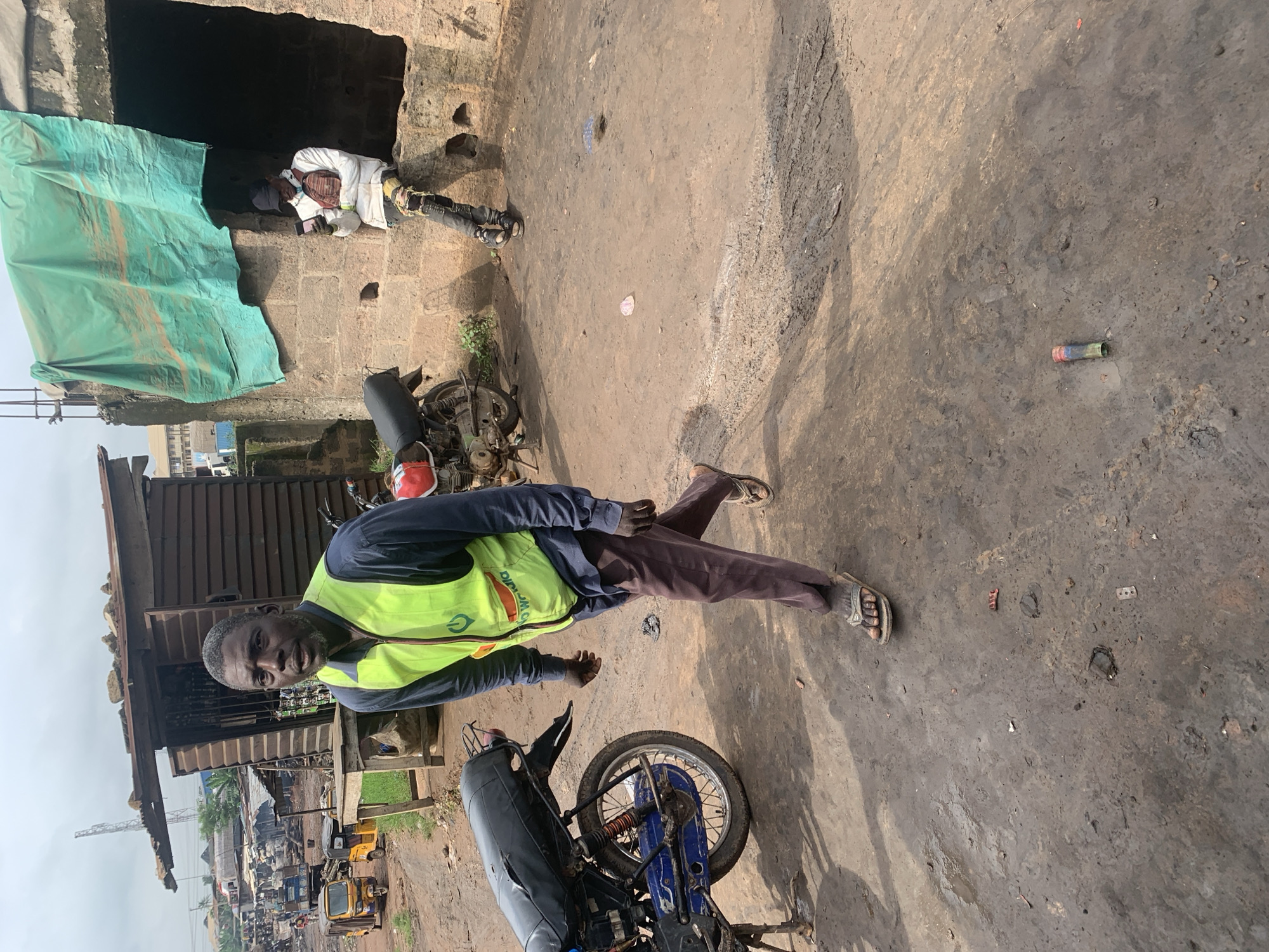 In the city of Shagamu, Nigeria, a man wearing a neon green vest over a dark long-sleeved shirt and brown pants walks across a dirt ground. He is wearing dark sandals. Behind him, a motorcycle is parked on the left side of the frame. Further in the background, there is a wooden structure with a metal roof and a green tarp hanging from it. Another person, wearing a white hooded jacket and dark pants, is sitting on a ledge on the right side of the frame, looking at their phone. They are wearing a dark cap and a brown cross-body bag. The environment appears to be an outdoor market or workshop, with various vehicles and structures visible in the distance. The ground is uneven and covered in dirt and debris.