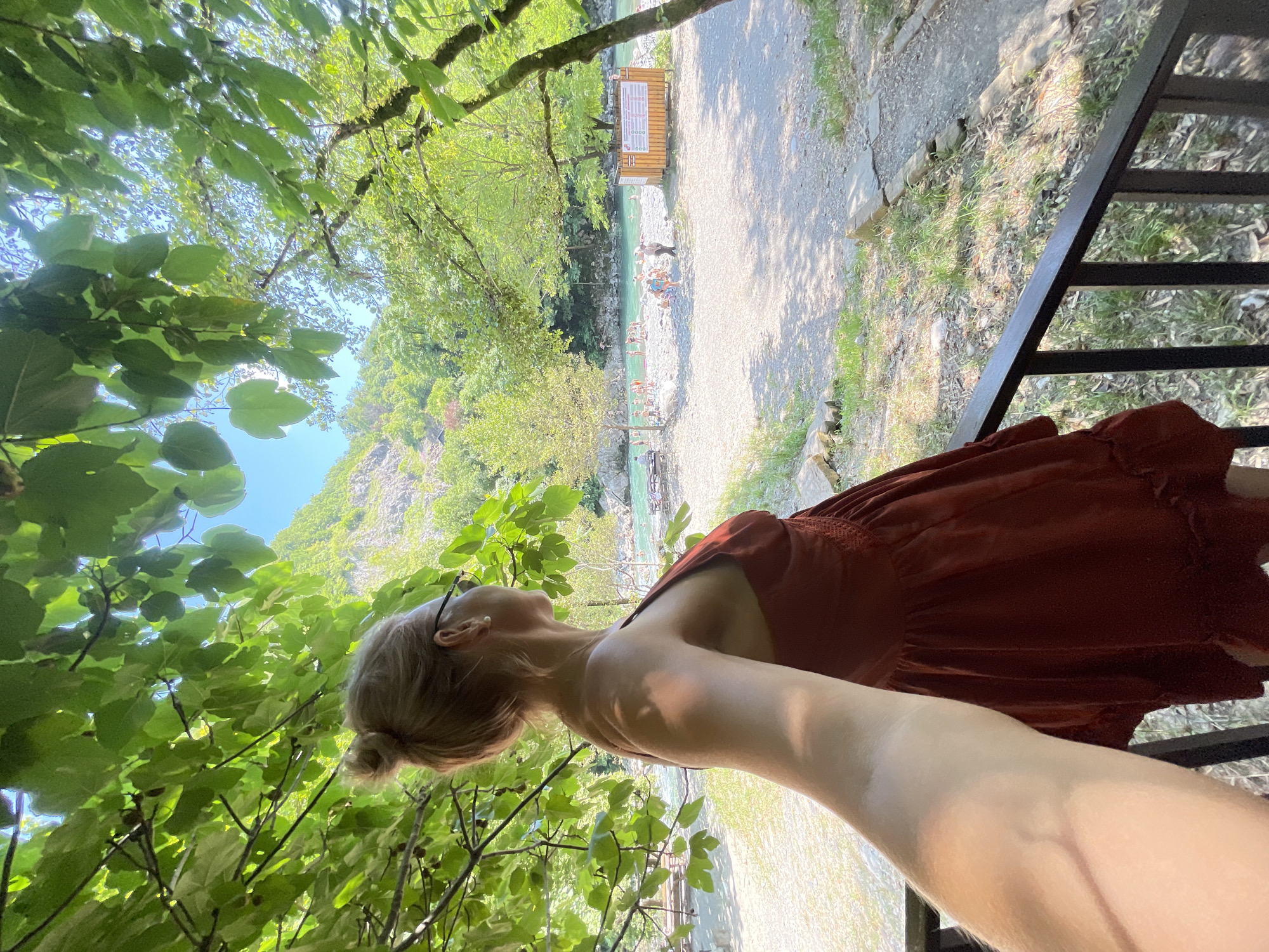 This is a selfie taken by a blonde woman with her hair in a bun, wearing a red dress and sunglasses, who is standing on a wooden platform overlooking a rocky riverbank with turquoise water and a wooded mountainside in the background. She is looking to her right, away from the camera. In the distance, several people are visible on the riverbank, some in the water. A wooden information board with text in a foreign language is present on the right side of the riverbank. The scene appears to be a natural outdoor recreational area, possibly a beach or swimming spot along a river, in a mountainous, forested region. The lighting suggests it is a sunny day. There is no direct indication of the location being specifically in Sochi, Russia.