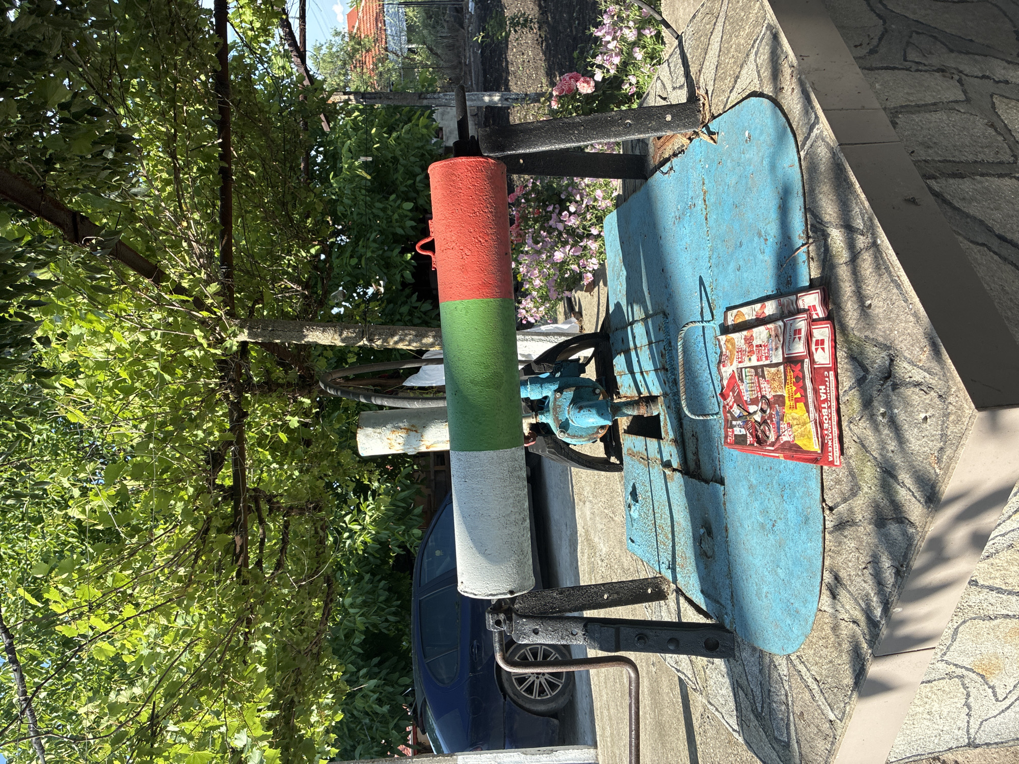 The image captures an outdoor scene in Radievo, Bulgaria, featuring a manually operated well or water pump in the foreground, painted in the colors of the Bulgarian flag (white, green, red). The pump mechanism, largely obscured, includes a blue metal base with visible rust and a dark, possibly cast-iron, support structure. A red folded flyer or advertisement, bearing the "Kaufland" logo and Cyrillic text, rests on the blue surface. A small, dark object, possibly a pair of spectacles or keys, lies on the flyer.
The well structure is positioned on a raised, grey-tiled platform with a textured, stone-like appearance. Strong sunlight casts distinct shadows across this platform and the pump.
In the mid-ground, a dense canopy of green grapevines or similar foliage extends overhead, supported by a metal framework, providing shade. To the left, a dark blue sedan (likely a Peugeot 407 model, identifiable by its wheel design) is parked on a paved area, partially visible behind the well. Further left, mature green trees form a backdrop. To the right, a garden bed features numerous pink flowering plants, likely roses, with tilled earth behind them. In the far background on the right, a portion of a building with a red-tiled roof is visible under a clear sky. No people are present in the frame.