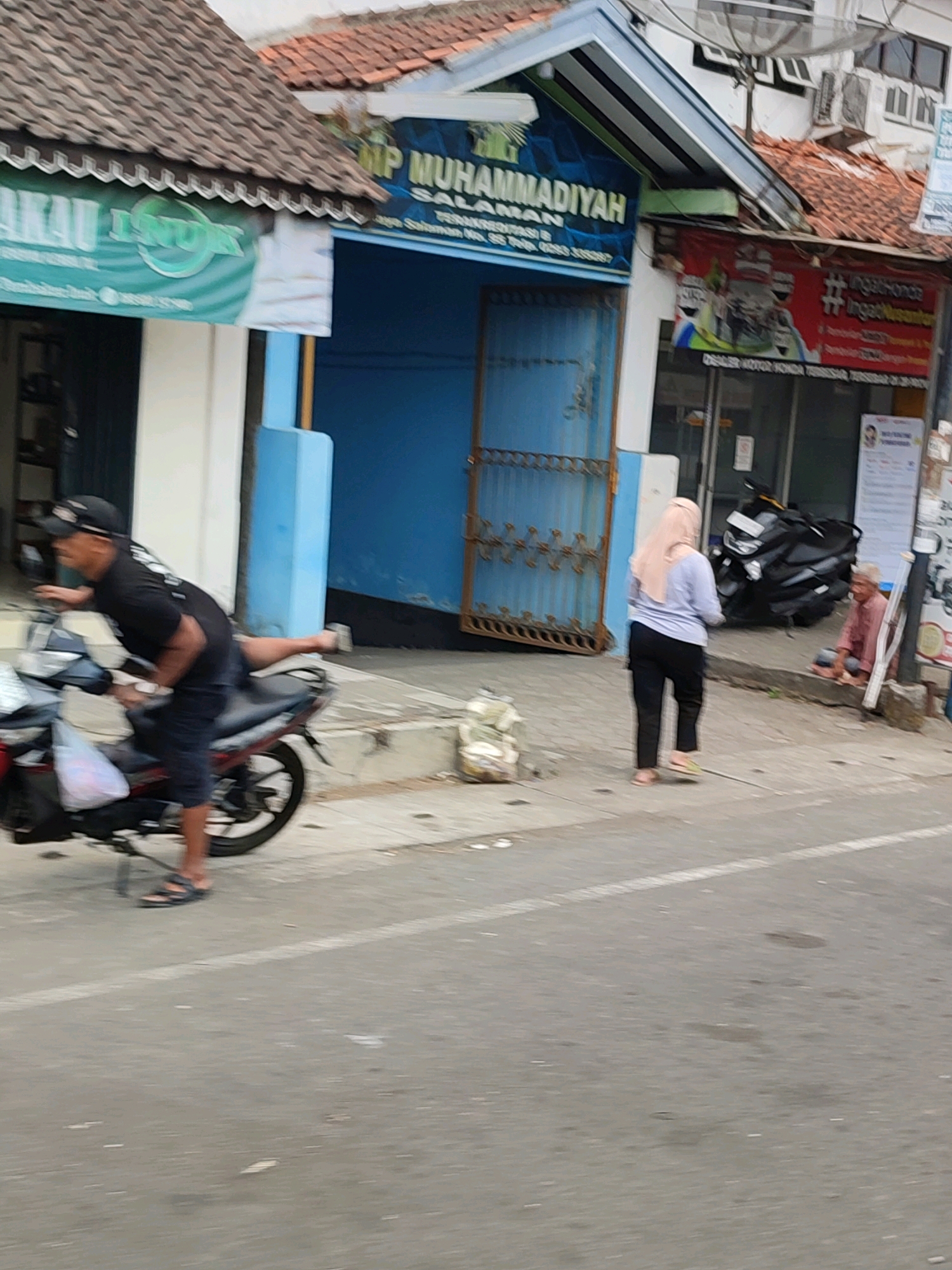 In the city of Salaman, Indonesia, a man wearing a black t-shirt and a cap rides a dark-colored motorcycle with a red frame, leaning to his left with his right leg extended backward. He is on a paved road with a white line. To his right, on the sidewalk, a woman in a light pink hijab and a light blue shirt with black pants walks away from the viewer. Behind her, a black motorcycle is parked. Further to the right, an elderly man with a pink shirt and a light-colored cloth sits on the ground next to a pole, appearing to be seeking alms. In the background, there is a building with a tiled roof and blue doors, bearing a sign that reads "SMP MUHAMMADIYAH SALAMAN". Another banner on the building shows "LINGKAR IND". The environment appears to be an urban street with various businesses and buildings visible.