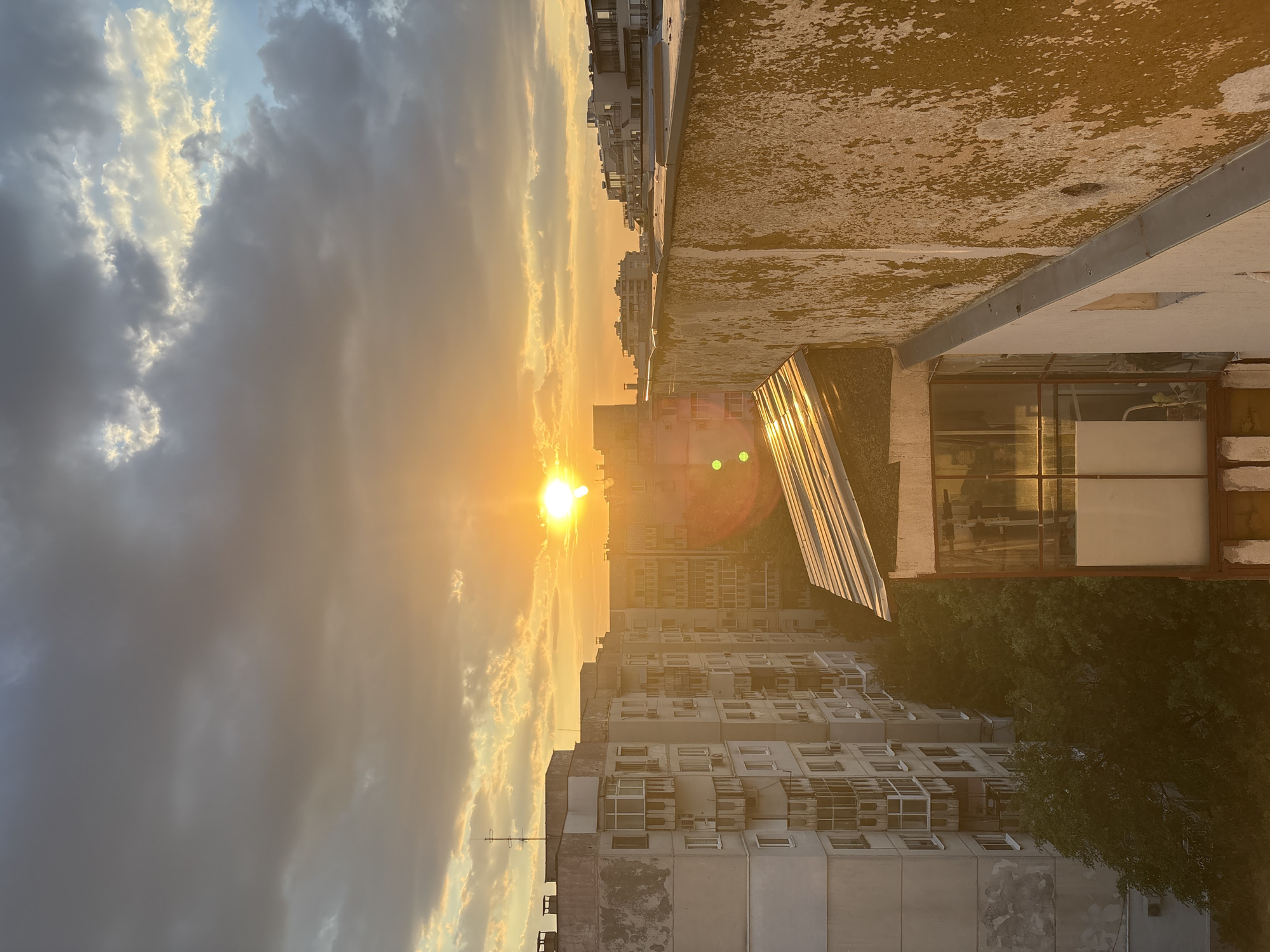 This is a vertical, medium-shot photograph taken from a rooftop at sunset in Plovdiv, Bulgaria. The sun is low in the sky, casting a warm, golden light that illuminates a densely populated urban scene. The sky is a mix of dramatic, dark clouds and bright, sunlit wisps, suggesting a dynamic weather pattern.
The foreground is dominated by a weathered rooftop, its surface appearing rough and uneven with patches of discoloration, possibly due to age and exposure. A reflective, angled structure, perhaps a skylight or solar panel, captures the sunset's glow, creating a bright glare.
Beyond the rooftop, a sprawling cityscape of apartment buildings stretches into the distance. The buildings are of varying heights, many featuring rows of windows and balconies adorned with air conditioning units. Some structures appear older, showing signs of wear and tear, while others seem more modern. Lush green trees are visible interspersed among the buildings, adding a touch of nature to the urban environment. The overall impression is one of a lived-in, bustling city at the end of the day. The golden hour lighting evokes a sense of warmth and perhaps nostalgia. No visible text is present in the image.