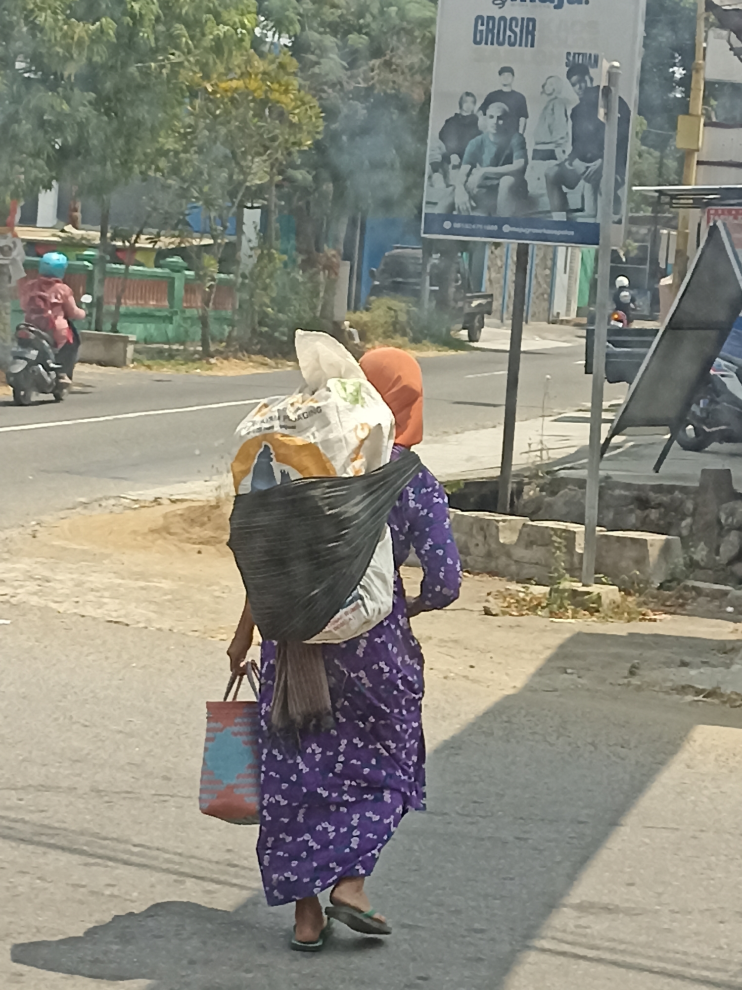 In Pulisen, Indonesia, a woman walks down a street carrying a large bundle on her back, secured with a dark fabric. She is wearing a purple patterned dress and an orange headscarf, with a patterned bag in her left hand. A billboard with an advertisement is visible in the background. The environment appears to be a sunny outdoor setting with some vegetation and other pedestrians and vehicles present.