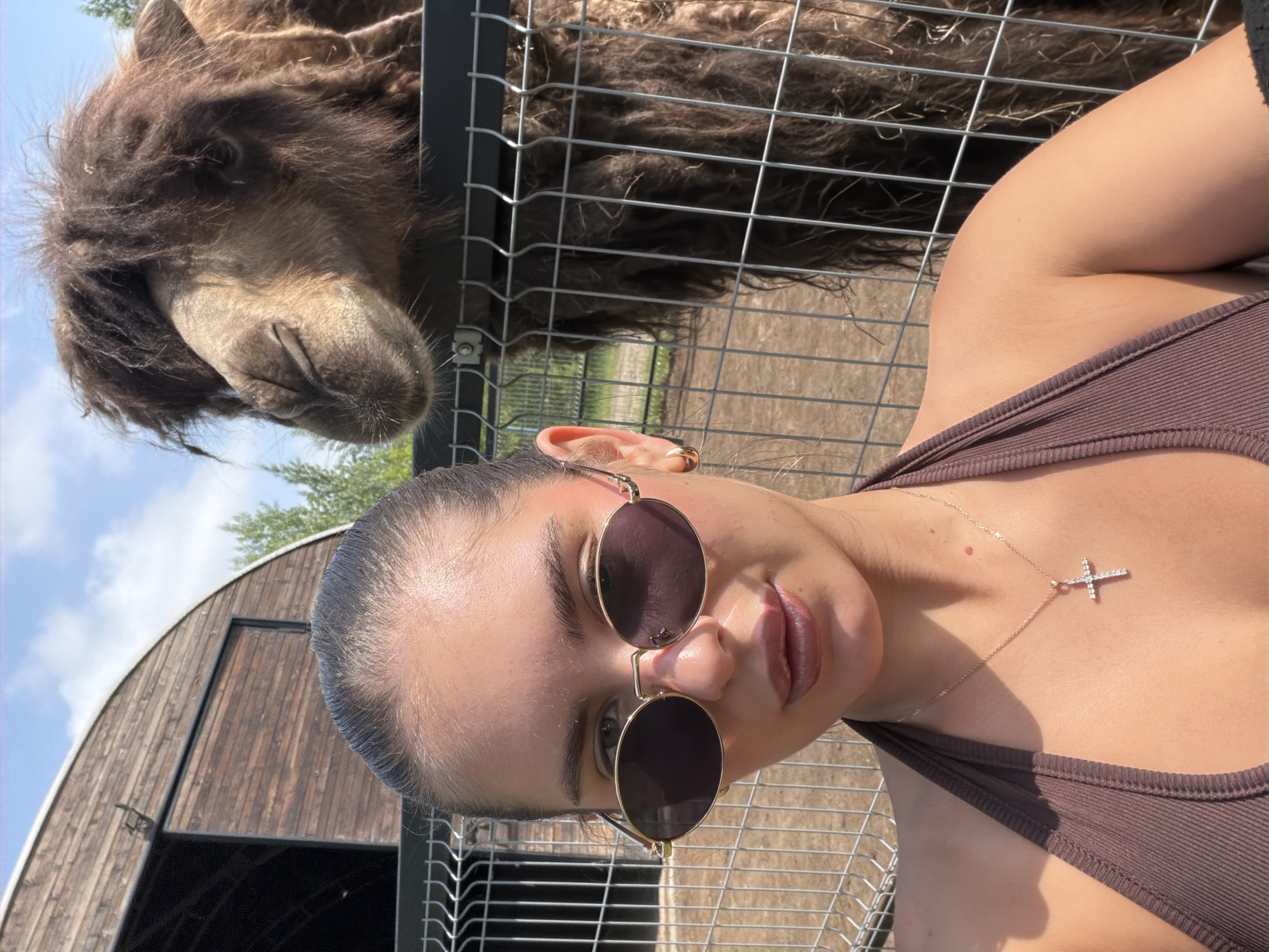 A young woman with dark hair pulled back is taking a selfie. She is wearing sunglasses and a brown halter top with a cross necklace. Behind her, a camel is visible, with its head over a fence. The setting appears to be an outdoor enclosure, possibly a zoo or sanctuary, with a wooden building and trees in the background. The lighting suggests it is daytime, with the sun casting shadows.