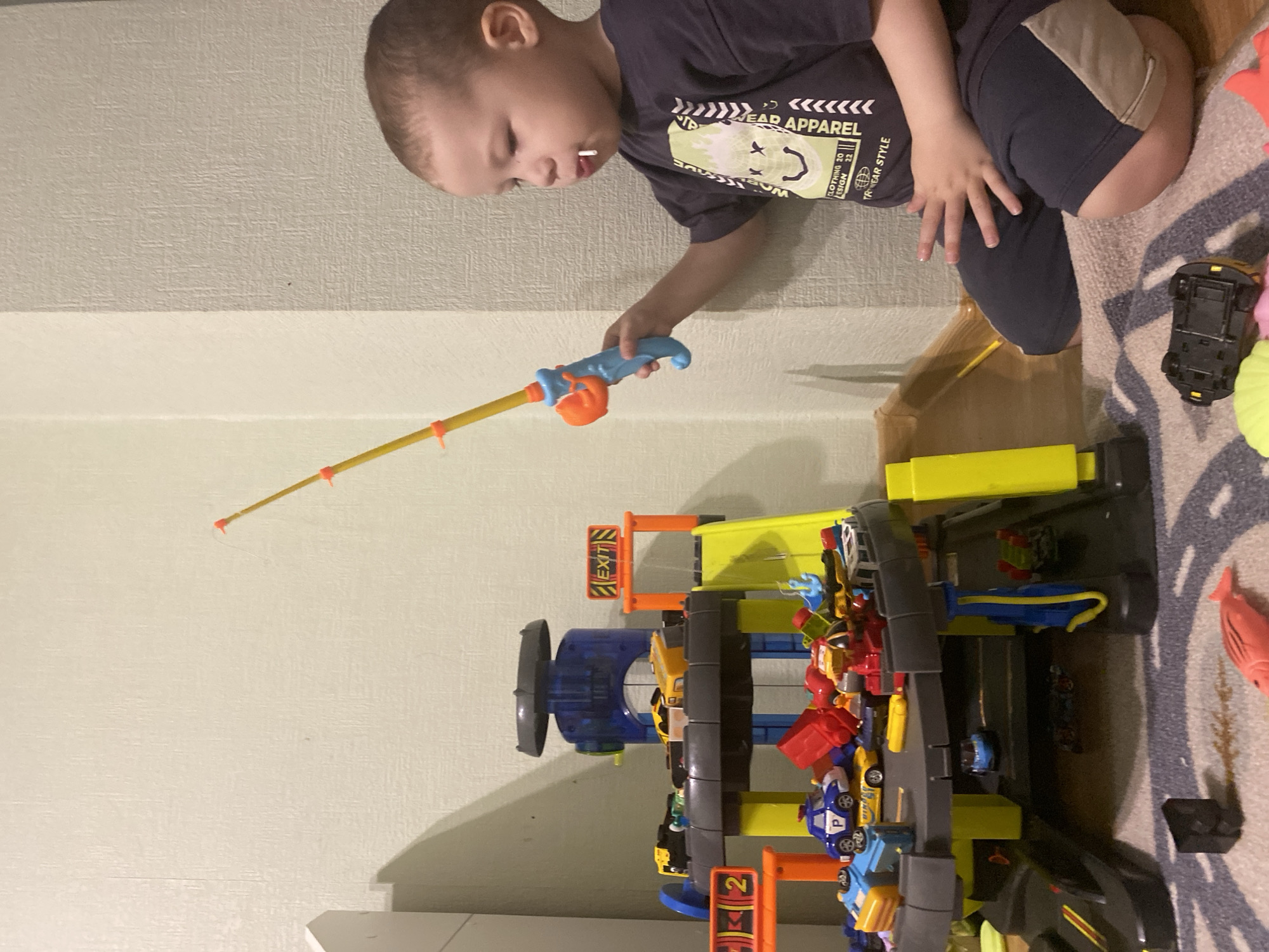 A young child is playing with a toy fishing rod and a multi-level toy car garage filled with toy cars. The child is kneeling near the wall, holding the fishing rod towards the garage. The garage is full of colorful toy cars. The background includes a textured wall and a patterned rug.
The location is not identifiable as Sochi, Russia based on the image.