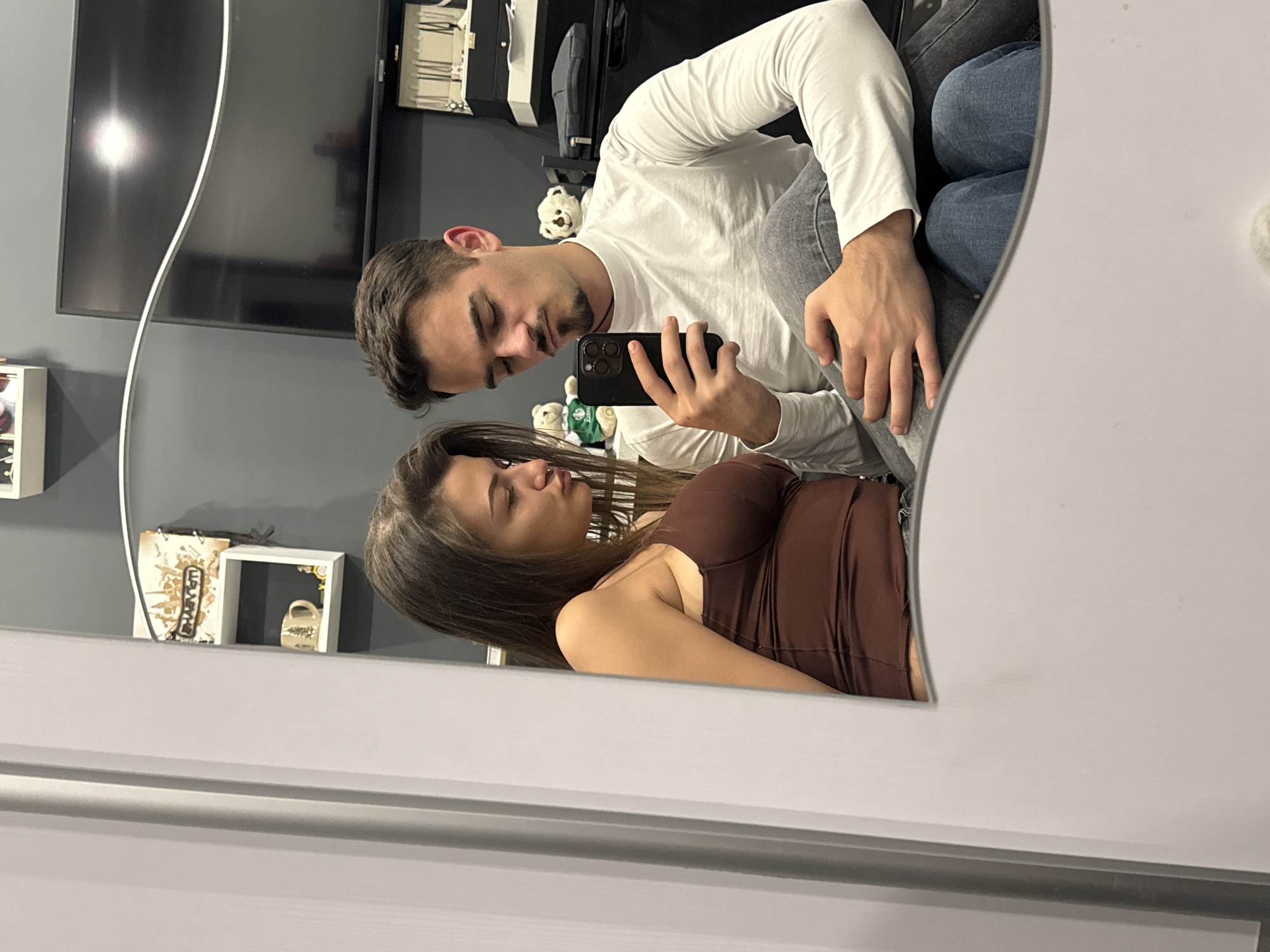 A photo shows a man and a woman reflected in a mirror. The man, wearing a white long-sleeved shirt and blue jeans, is holding a black smartphone and looking at it. The woman is wearing a brown dress. The background includes a gray wall, a television, and various objects on shelves. No location can be determined.