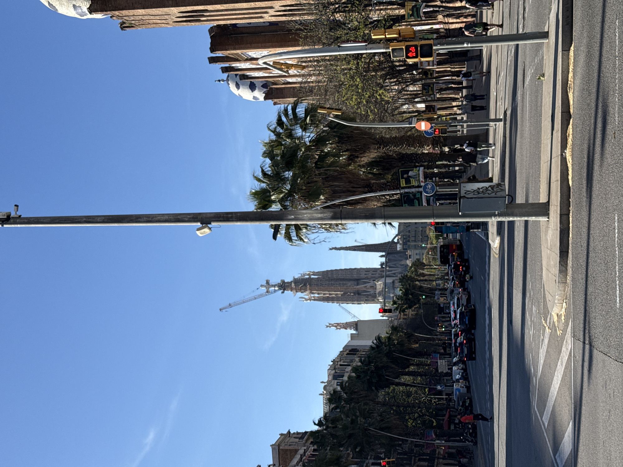 The image shows a street scene in Barcelona, Spain. Sagrada Familia is visible in the distance, partly obscured by palm trees. A crane is positioned near the basilica. The street has pedestrian crossings and traffic. People are walking on the sidewalk, and cars are lined up on the street. Traffic lights are visible, showing red. Buildings line the street, and the sky is blue.