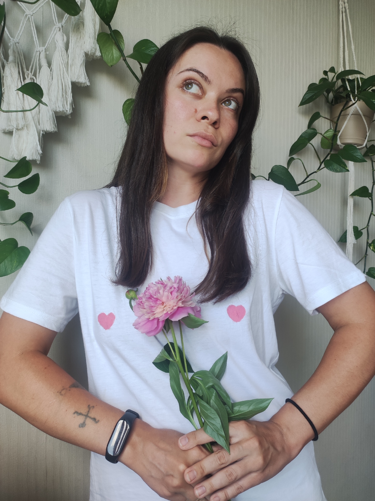 A woman with long, dark hair is holding a pink peony and looking upwards with a thoughtful expression. She is wearing a white t-shirt with two small pink hearts printed on it and a black fitness tracker on her left wrist. She has a tattoo on her left forearm. The background consists of a textured light-colored wall, a macrame wall hanging with white tassels on the left, and a potted plant with green leaves hanging from the ceiling on the right. The lighting appears to be natural and soft. The scene is indoors, in what could be a residential setting. There is no indication that this image is located in Chaikovsky, Russia.