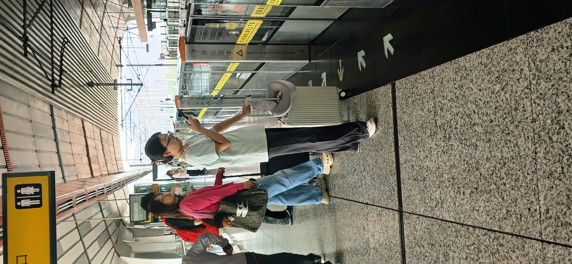 The image shows a group of people waiting at a subway station in Chongqing High-tech Zone, China. A woman wearing glasses and a light green shirt is looking at her phone, while standing next to a silver suitcase. Beside her, another woman in a pink shirt is also looking at her phone, holding a leopard-print bag. In the background, there are subway doors with caution signs in Chinese and English. The floor is gray with black guiding arrows. A yellow sign for restrooms is also visible.