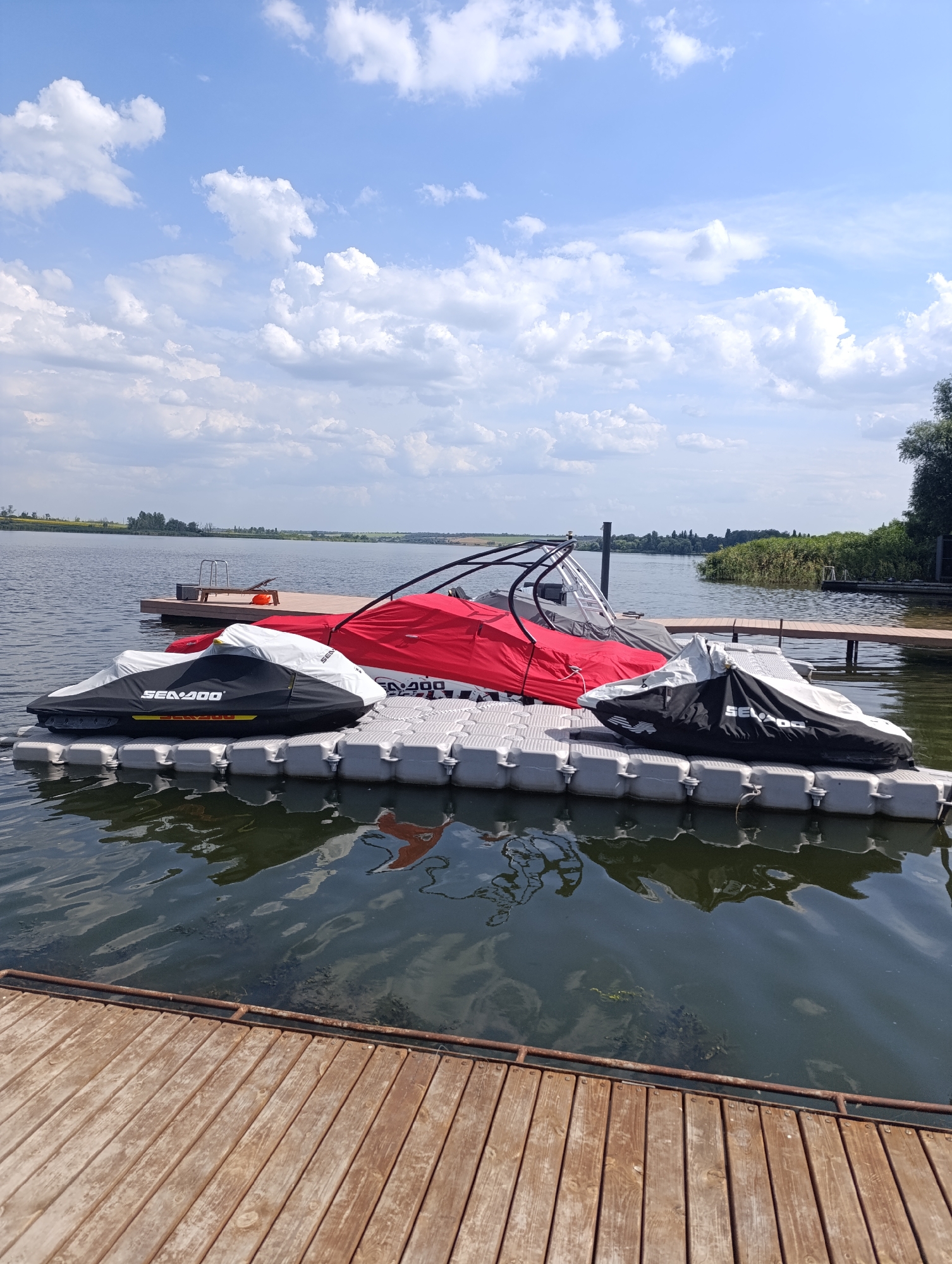 This image depicts a waterfront scene in Bulatselivka, Ukraine, under a partly cloudy sky. In the foreground, a wooden dock extends into a calm body of water. Moored to a modular floating dock system are two personal watercraft, each covered with a protective tarp. The watercraft on the left is covered with a black and white tarp bearing the "Sea-Doo" logo. To the right of this, another personal watercraft is partially visible, covered by a red tarp. Further to the right, a third personal watercraft is covered by a black and white tarp, also with a visible "Sea-Doo" logo. Behind the floating dock, a larger, more permanent wooden dock is visible, with a ladder and seating area. The background shows a treeline and vegetation along the shoreline, with fields and a hazy sky further in the distance. No people are visible in the image.