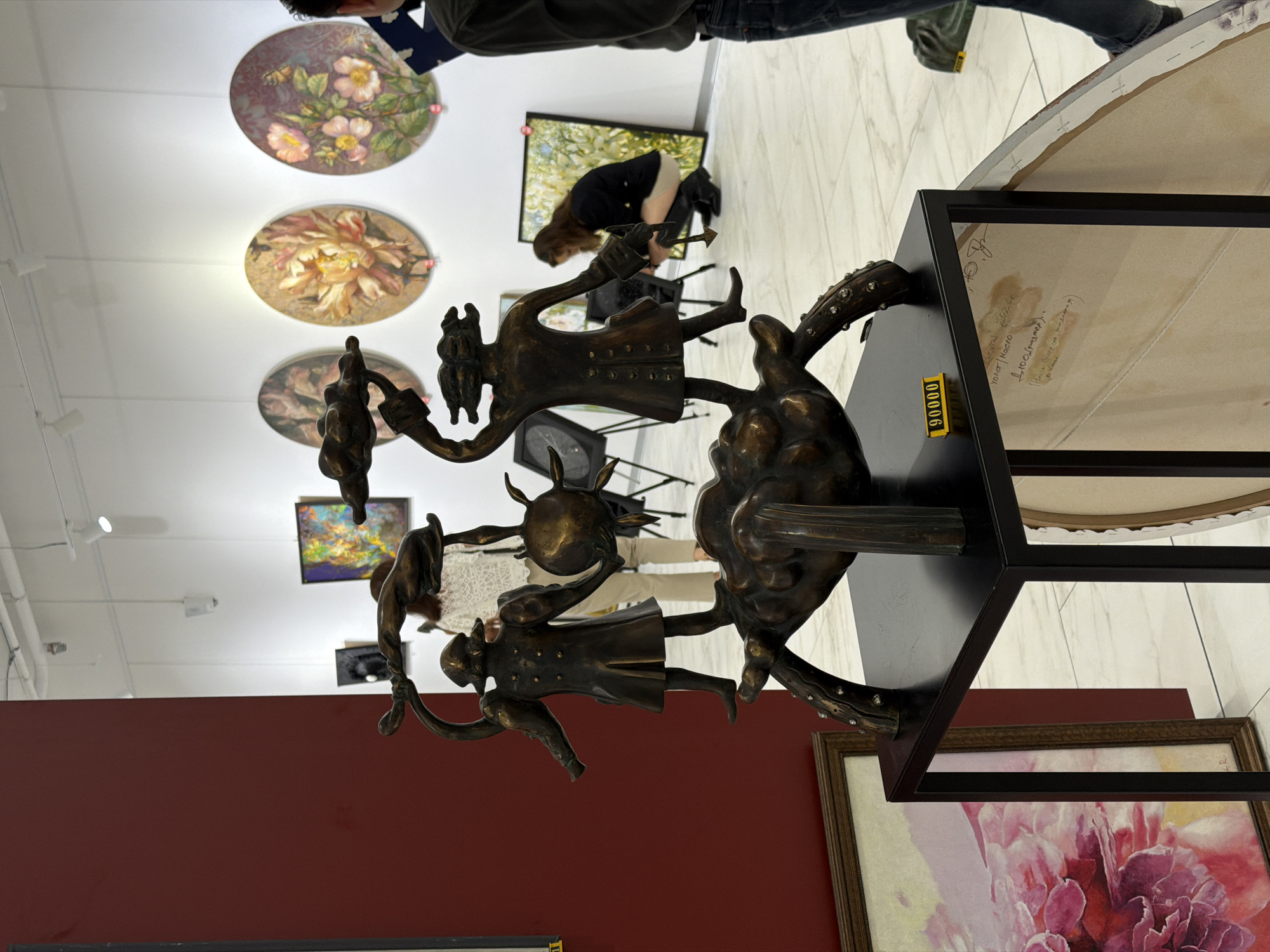 This is a medium shot of a bronze sculpture displayed in an art gallery, likely located in Minsk, Belarus, given the Cyrillic writing on the back of a canvas in the foreground. The sculpture depicts two figures standing on a cluster of what appears to be eggs or pods, holding objects above their heads. One figure is on the left, wearing a coat with large puffed sleeves and buttons, and the other is on the right, similarly attired. Both figures have stylized, almost mask-like faces. The sculpture is dark bronze and appears to be antique or made to look so.
The sculpture is placed on a black metal pedestal. A yellow price tag with the number "90000" is visible on the front of the pedestal. To the left of the pedestal, leaning against a dark red wall, is a framed painting of a large pink flower. To the right of the pedestal, a partially visible canvas is propped up, with what looks like handwritten notes in Cyrillic on its back.
In the background, the gallery walls are white, adorned with various artworks. Three circular paintings featuring floral motifs are prominently displayed on the back wall. Further back, other pieces of art are visible, some framed and some on easels. There are several people in the background, blurred and out of focus. On the right side, a person in dark clothing is seen from the waist down, standing. A woman in a black jacket and light-colored skirt is crouching down to examine a painting on an easel. Another person, wearing a white lace top and beige pants, is visible standing behind the sculpture.
The floor of the gallery is tiled with large white marble-like tiles. The lighting in the gallery appears to be artificial, with track lights visible on the ceiling. The overall impression is that of a well-lit, organized art exhibition space.
