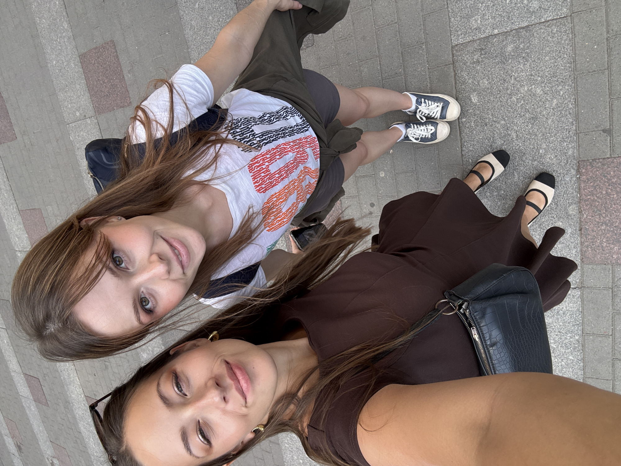 A high-angle selfie captures two young adult women standing on a paved sidewalk in Saint Petersburg, Russia, under bright daylight. The woman on the left, holding the camera, has long dark brown hair, gold hoop earrings, and sunglasses perched on her head. She wears a dark brown short-sleeved dress and black and beige two-tone Mary Jane style flats. A small black textured shoulder bag is visible on her left side. She winks with her right eye and smiles faintly. The woman on the right has medium brown hair with bangs and wears a white t-shirt featuring the word "BOLD" in prominent orange text and additional black text below it. She is dressed in dark grey shorts with an olive green jacket tied around her waist. She wears blue canvas low-top sneakers with white laces and white socks. A dark backpack is visible on her back. Both women are smiling towards the camera. The sidewalk is paved with a pattern of rectangular grey and reddish-brown concrete blocks. The specific location within Saint Petersburg cannot be determined from visual cues.