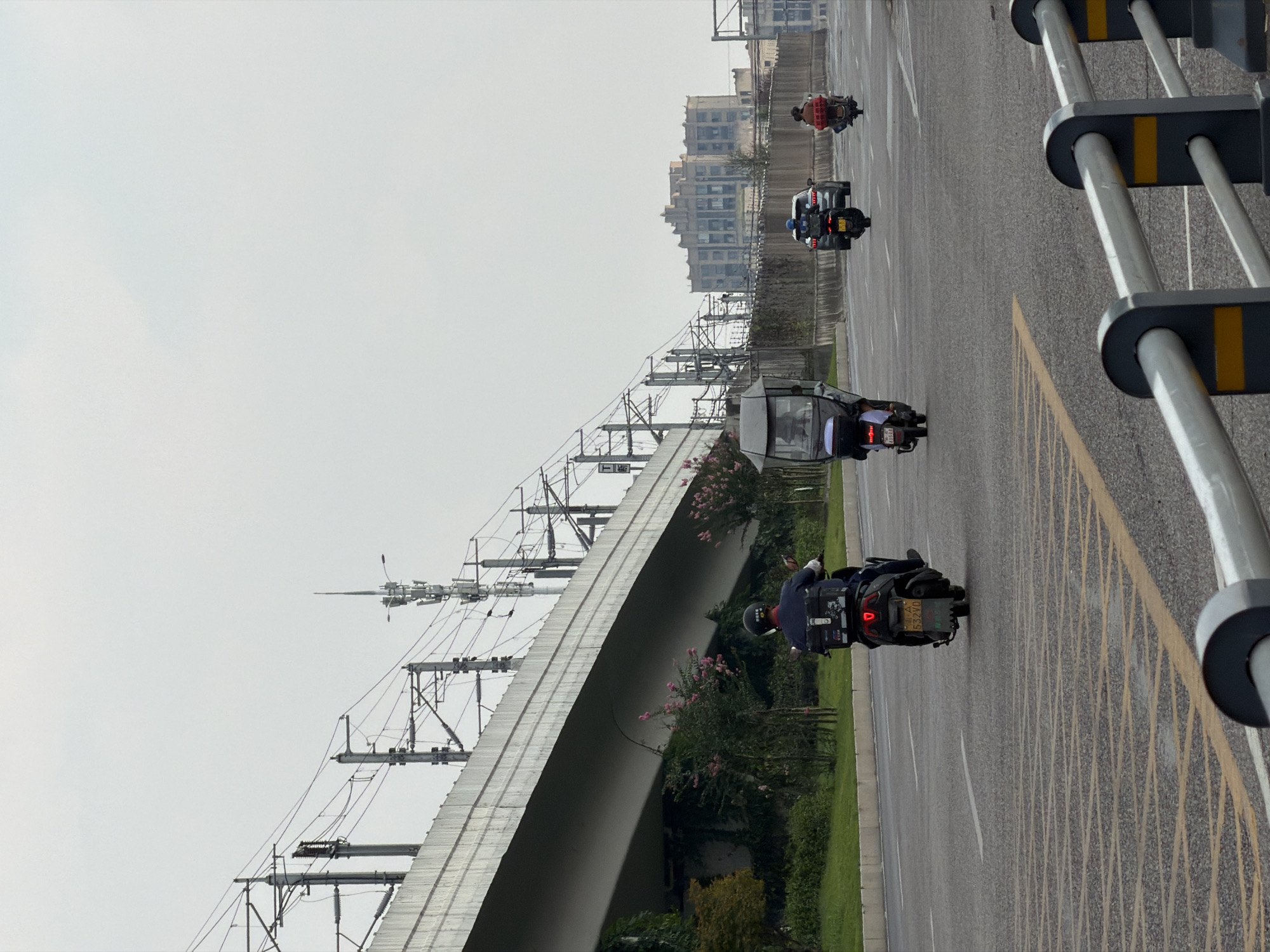 In Jiangjin, China, a street scene unfolds under an overcast sky, dominated by a wide elevated roadway or overpass constructed from concrete. This structure is supported by numerous pylons and is bordered by railway overhead lines and communication towers. Below the overpass, a strip of green grass and foliage lines the road. On the asphalt road, several vehicles are in motion. In the foreground, a person on a scooter, wearing a dark blue uniform and helmet, is riding away from the camera, their scooter equipped with a black storage box on the back and a yellow license plate. Further down the road, another scooter with a covered canopy is visible, with a person inside. In the middle distance, a silver car is traveling, followed by a person on a red scooter. On the right side of the road, another scooter is seen from behind. In the background, a large, multi-story building with a distinctive architectural style is visible, and more buildings are scattered further into the distance. A metal guardrail with yellow accents runs along the right side of the road in the foreground.