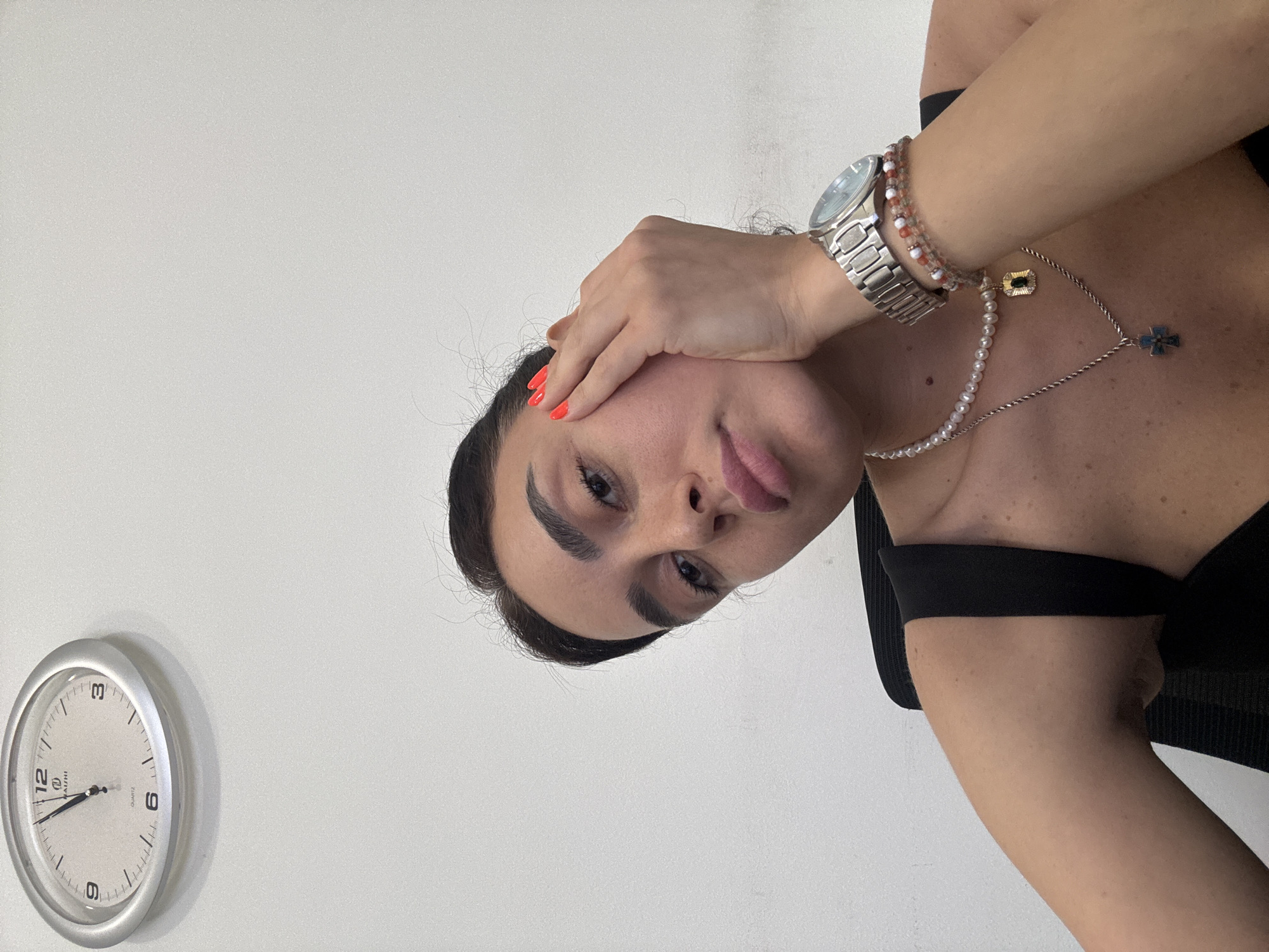 The image shows a close-up of a woman with dark hair, fair skin, and striking eyebrows. She is wearing a black top, pearl necklace with a gold pendant, and a silver wristwatch with multiple bracelets. Her right hand is touching her face, and her nails are painted a bright orange. She has a relaxed, pensive expression. In the background, a silver analog clock with a white face is mounted on a white wall. The clock's hands indicate approximately 12:55. The overall scene appears to be an indoor setting, likely an office or home, with a plain white background. The image does not provide any specific visual cues to determine its location within Tbilisi, Georgia.