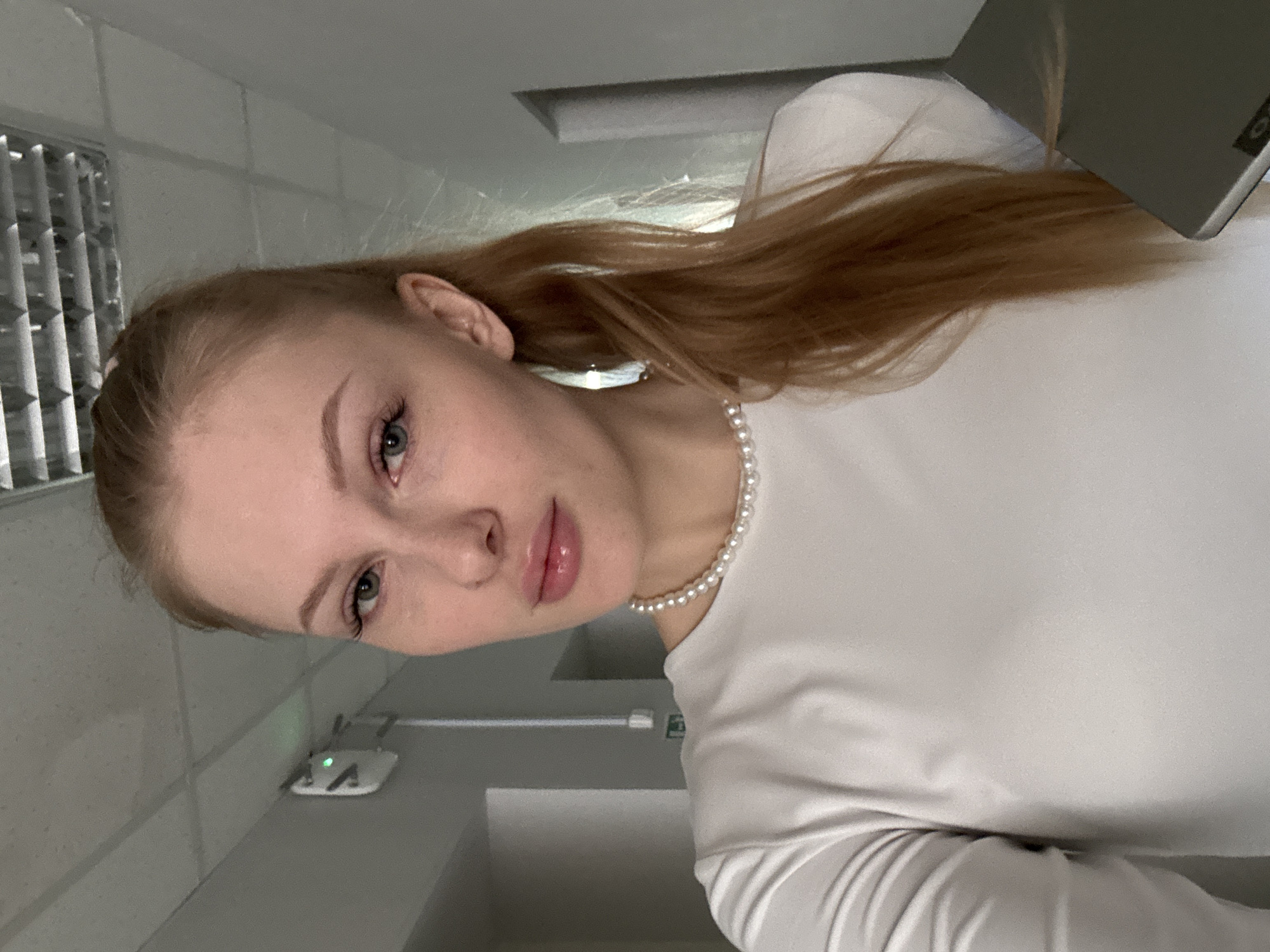 This vertically oriented photo features a young woman posing for a portrait in an indoor setting, likely in Perm, Russia. She has fair skin, light-colored eyes (appearing blue or green), and long, reddish-blonde hair that cascades over her shoulder. Her makeup is subtle, with defined eyebrows and glossy pink lips. She wears a simple white long-sleeved top and a pearl necklace.
The background suggests an institutional or public building environment, with grey walls and white ceiling elements. In the upper left, a metallic ventilation grate is visible. Below the woman's face, on a shelf or ledge, rests a small white electronic device with green indicator lights, possibly a Wi-Fi router. Partially visible in the upper right corner is a dark-colored laptop, with the "Lenovo" logo faintly discernible on its lid. The overall lighting is artificial and somewhat dim, typical of an indoor setting. The woman looks directly towards the camera with a calm and neutral expression, indicating a posed shot or selfie. No specific activity beyond posing is captured.
