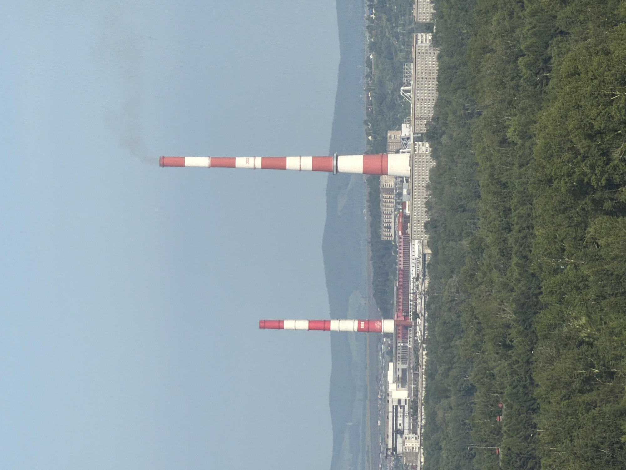 This media file shows a wide shot of an industrial area situated next to a densely forested landscape and a body of water. The image is vertically oriented. Two prominent industrial chimneys, painted with alternating red and white stripes, are the main subjects. The taller chimney, on the left, is emitting a plume of dark smoke into the clear, light blue sky, suggesting an active industrial process. The second, shorter chimney is positioned further to the right and appears to be idle, with no visible smoke.
In the background, a city skyline with numerous tall, uniform buildings can be seen stretching along the edge of the forest and water. These buildings suggest a populated urban area adjacent to the industrial zone. The forest is lush and green, indicating a temperate season, likely summer or late spring. The water is calm and reflects the sky, providing a serene contrast to the industrial elements.
The overall scene depicts a juxtaposition of heavy industry and natural beauty, typical of many urbanized areas in Russia. The clear sky and bright lighting suggest it is daytime, likely mid-morning or afternoon. There is no visible text in the image, apart from what appears to be markings on the taller chimney, possibly a serial number or operational code, but it is not legible. The image evokes a sense of industrial activity within a developing landscape.