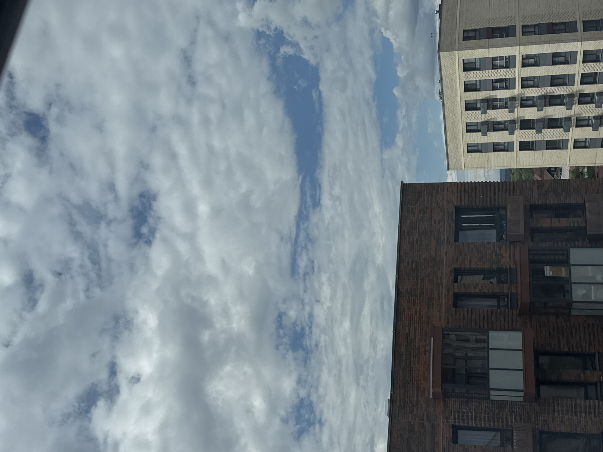 A daytime, eye-level, outdoor, medium shot captures a view of two modern apartment buildings against a cloudy sky. The building on the left is constructed with dark reddish-brown bricks and features glass-enclosed balconies with frosted panels. The building on the right is lighter in color, appearing to be made of beige brick or concrete, with numerous dark-framed windows and small, dark balconies. The sky is filled with a mix of white and gray clouds, with patches of blue sky visible. The lower portion of the image shows the tops of the buildings, indicating they are tall structures. No people are visible in the image. The environment appears to be urban, with the buildings suggesting a residential area.