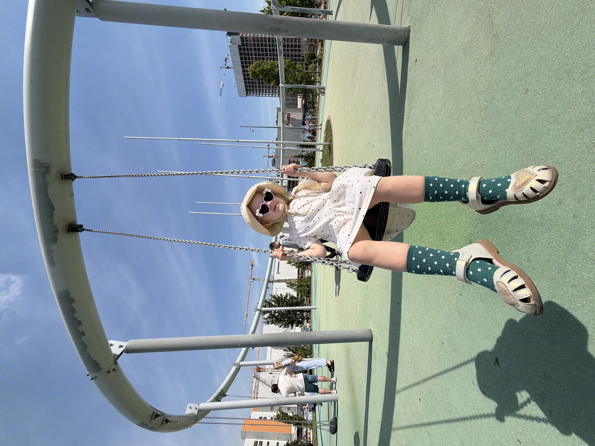 This image presents a full-length view of a young girl actively swinging on a modern playground structure under a clear, bright blue sky with minimal cloud cover, on a sunny day.
The central subject is a Caucasian girl, approximately 5-7 years old, captured mid-swing. She is wearing a light-colored (possibly white or cream) short-sleeved dress with small dark polka dots. Her head is adorned with a light-colored sun hat, and she wears heart-shaped sunglasses with decorative frames, obscuring her eyes. Her light-colored hair, possibly blonde, is visible beneath the hat, styled with two small braids. Her feet are clad in dark green socks with white polka dots and light-colored (beige or cream) open-toed sandals featuring a heart-shaped detail on the front. She holds onto the metal chains of the swing with both hands.
The swing set is an integral part of a contemporary, large-scale, modular playground structure. It consists of robust, light-gray, curved metal beams supported by vertical cylindrical posts, indicating a public park or recreational area. The swing seat itself is black. The ground beneath the playground is covered with a uniform light green, textured, soft-surface material, likely rubberized, designed for safety. Distinct shadows are cast on this surface by the girl, the swing, and the support columns, indicating strong sunlight from a high angle.
In the mid-ground and background, other individuals, including adults and children, are visible on the playground, some standing, some walking. To the left, an adult male in a white t-shirt and blue shorts converses with an adult female in a light-colored jumpsuit. The broader background reveals a modern urban landscape with large buildings. A prominent shopping center building displays the Cyrillic text "АУШАН" (Auchan) with its recognizable orange 'A' logo, and "АВИАПАРК" (Aviapark) is also discernible on an adjacent structure. To the right, a tall, dark-colored multi-story building is under construction, with its exposed framework visible, accompanied by multiple construction cranes against the skyline. These features indicate a location within a developed urban or suburban area, identified as being in the city of Khimki, Russia.
