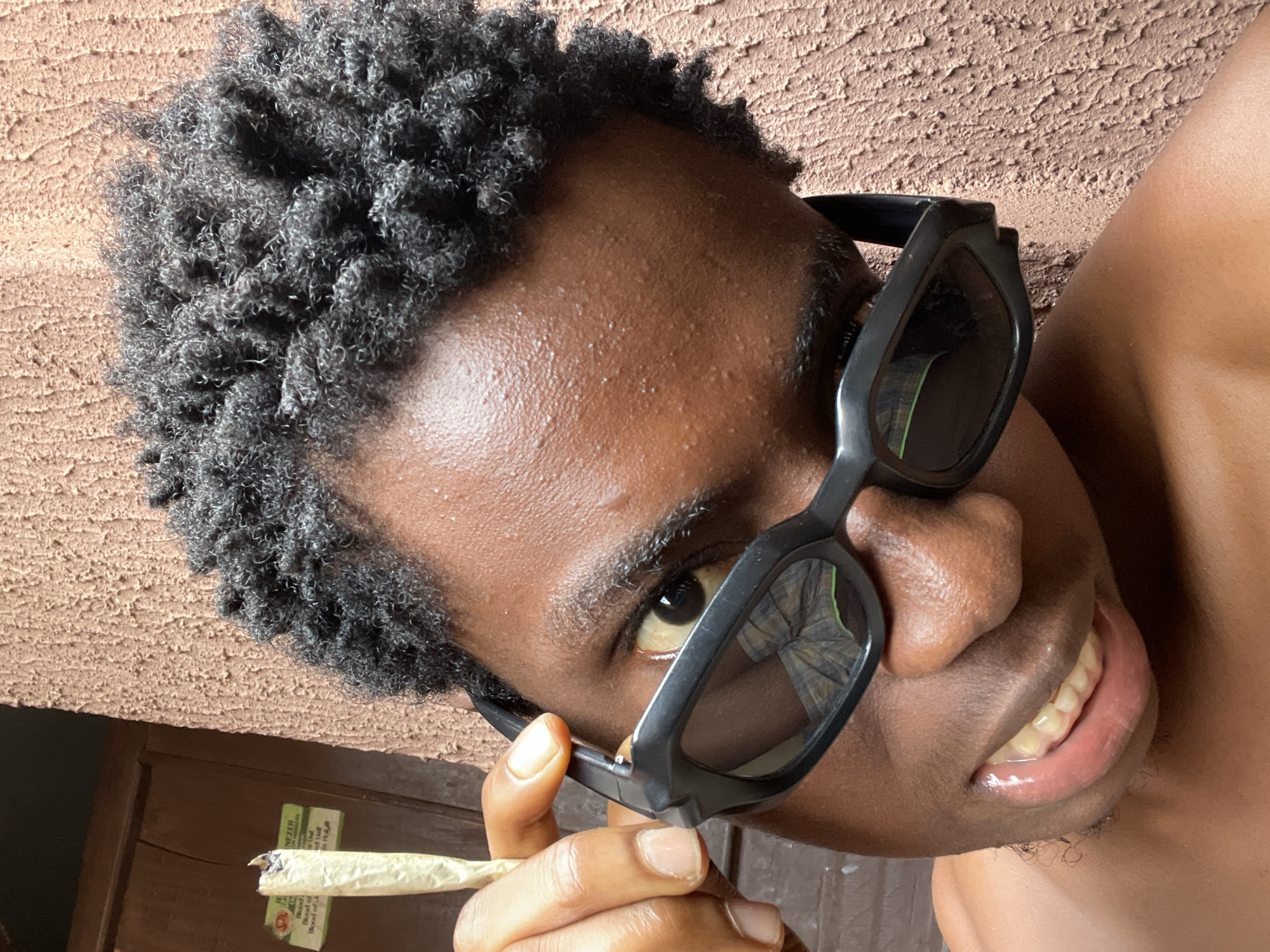 This media file is a close-up, rotated portrait of a young Black individual wearing black sunglasses and holding a lit cigarette in their right hand. The subject's dark, curly hair is visible, as are their forehead, eyes, nose, and mouth. The skin appears to be slightly sweaty. The background consists of a textured, light brown wall, and a darker wooden surface with some visible green text on a white label in the lower left corner. The lighting suggests it is daytime. The overall mood appears casual and relaxed.
Visible text includes:
- "NEZER"
- "PREMIUM"
- "BIKE OF"
- "BIKE OF A"