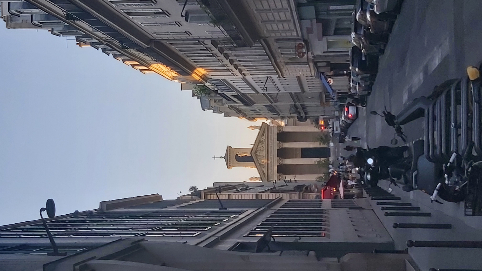 This is a vertical, eye-level, outdoor photograph taken on a street in Paris, France, during the late afternoon or early evening, as indicated by the warm, low-angled sunlight illuminating the right side of the buildings and the clear sky. The perspective looks down a street lined with tall, multi-story Parisian buildings.
On the left, a building with a modern facade features large windows with dark frames, reflecting the sky. A horizontal lamp fixture extends from the building.
On the right, a row of traditional Parisian buildings with mansard roofs and balconies runs along the street. Sunlight casts a warm glow on their upper floors and roofs. The street is lined with parked cars and several scooters or motorcycles are visible in the foreground on the left.
In the distance, at the end of the street, stands a classical-style building with a portico supported by large columns and topped with a pediment. A cross is visible on a tower above the pediment, suggesting it is a church or similar public building. Several small statues adorn the facade of this building.
People are visible walking on the sidewalks and crossing the street. Some are gathered near a cafe with red umbrellas on the left side of the street. The overall scene depicts a bustling urban environment in Paris, likely a residential and commercial area.