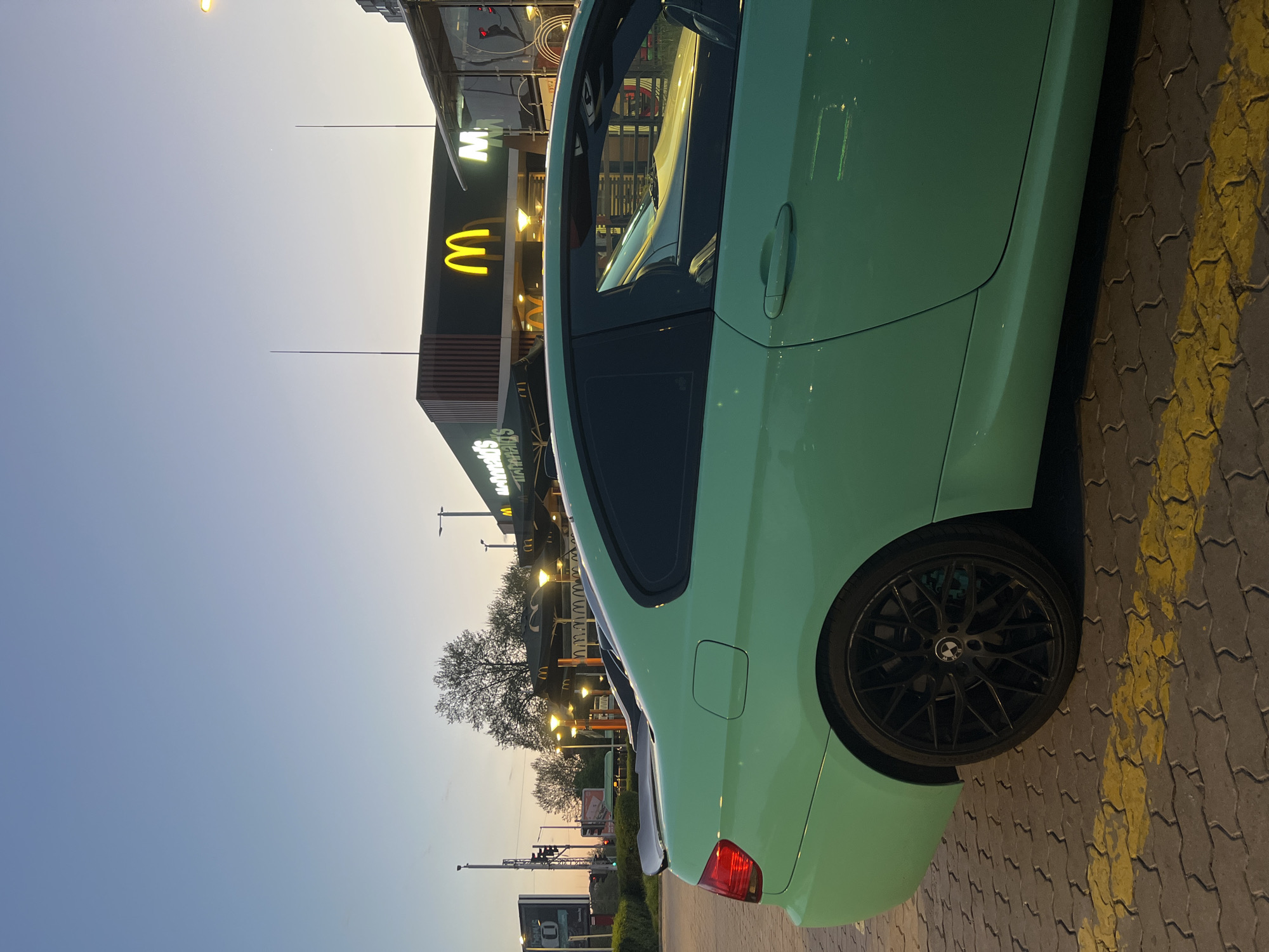 The image captures a distinctively colored car parked at dusk outside a McDonald's restaurant in Plovdiv, Bulgaria.
The primary subject is a light mint green or teal-colored car, likely a BMW judging by the logo on its black multi-spoke wheel. The car is seen from its rear-left side, showcasing its sleek bodywork and the red rear taillight. Reflections of the surrounding artificial lights are visible on its smooth surface. The car is parked on a paved area with interlocking grey/brown bricks, marked with irregular yellow painted lines on the ground to the right.
In the background, a modern McDonald's building stands prominently, illuminated by its own lighting. The iconic yellow "M" logo is brightly lit, standing out against the dark facade. Below the main "M," the full "McDonald's" text is also visible, along with other smaller, less distinct text that might indicate a drive-thru or service offerings. Several streetlights are visible, adding to the evening glow. Behind the restaurant, bare trees can be seen silhouetted against the sky.
The scene depicts the transition from day to night, with the sky displaying a gradient of light blue fading into a warm yellow-orange hue near the horizon. This twilight atmosphere suggests the time of day is either late evening or early morning, but the illuminated signs point towards evening. No people are visible, and the overall activity is static, showing a parked car in a commercial setting. The image evokes a sense of urban twilight, possibly after a quick stop for food.