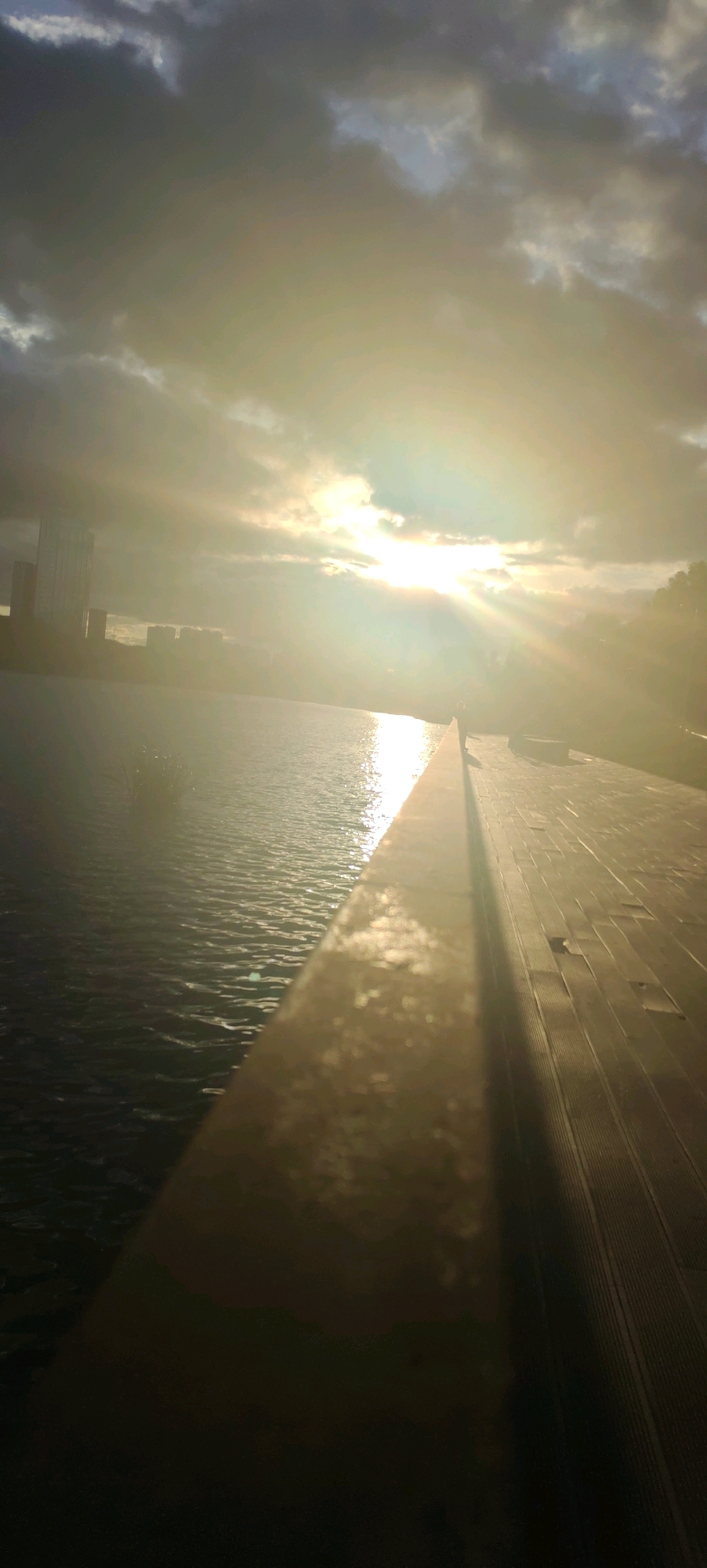 The image depicts a waterfront scene at dusk with a bright sun setting behind dramatic clouds. The sun's rays pierce through the clouds, creating a lens flare effect and casting a strong light across the water and a wooden boardwalk. On the left side of the image, a body of water is visible, with ripples on its surface reflecting the light. In the background, silhouetted buildings suggest an urban environment. On the right, the wooden boardwalk stretches into the distance, with its planks clearly visible. A single figure is standing on the boardwalk, silhouetted against the bright light. The overall atmosphere is serene and dramatic, characteristic of a sunset over water. The scene is consistent with a location along the Moskva River in Moscow, Russia, possibly a developed embankment area with modern architecture visible in the distance.