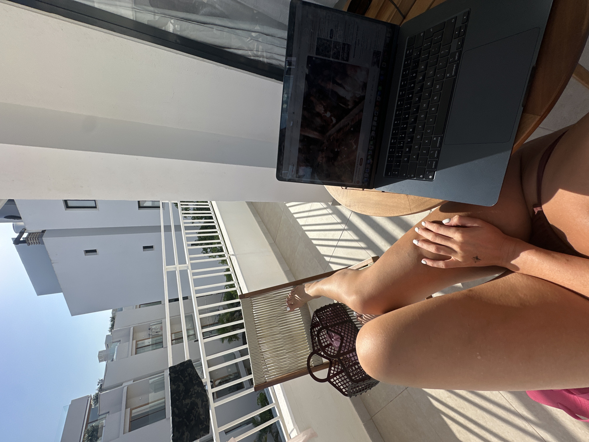 The image captures a first-person perspective from a sunny balcony in Eivissa, Spain, showing a person's legs and a laptop. The individual, likely female, is seated on a woven chair, with tanned legs crossed and painted white fingernails visible on one hand. A small star tattoo is visible on the back of the hand.
A dark gray laptop, resembling a MacBook, rests open on a small round wooden table to the right. Its screen displays what appears to be a video streaming platform, possibly YouTube, with various thumbnails and text that are difficult to read due to glare. The keyboard is visible, showing standard QWERTY layout and "Command" and "Control" keys. Strong sunlight casts distinct striped shadows across the balcony's tiled floor and the person's legs, suggesting it is mid-morning or late afternoon on a clear day.
Next to the person's feet, a dark brown, intricately woven basket is placed on the floor, and a pink fabric item is partially visible beneath the legs. The balcony features a clean white balustrade. In the background, modern, minimalist white buildings with flat roofs and rectangular windows are visible under a bright, clear blue sky. The architecture suggests a resort or residential area. The overall scene conveys a sense of relaxed work or leisure in a warm, sunny environment.