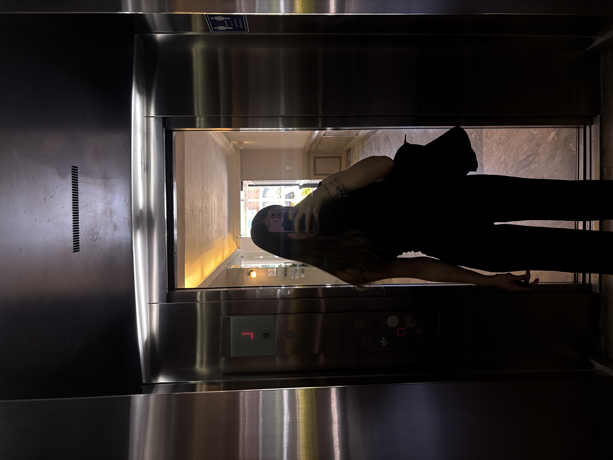 The image depicts a person taking a selfie in an elevator. The elevator has stainless steel walls and is on the 1st floor ("1" illuminated in red on the control panel). The control panel has buttons. The person is silhouetted, wearing dark clothing and holding a phone. The elevator door is partially open, revealing a hallway with light-colored walls. The location is Mexico City, Mexico.