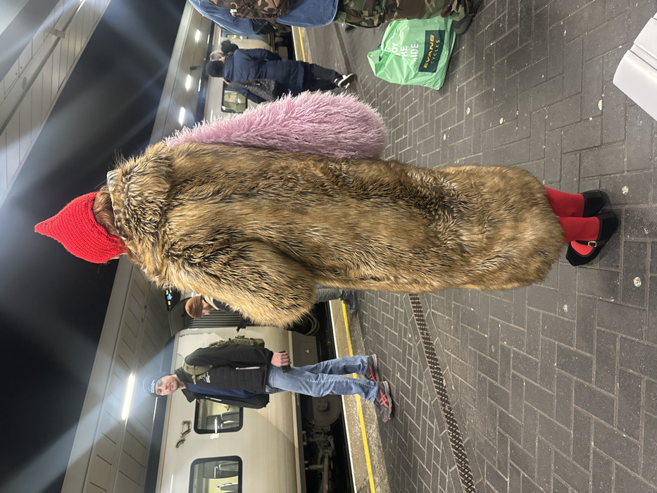 A person with a red pointy hat and a large fur coat stands at a train station. The coat has a large, fluffy pink detail on the back. The person wears bright red tights and black shoes. Another person in jeans, a blue beanie, and a black vest stands nearby. A train is visible in the background, and a green shopping bag sits on the platform. The platform is paved with dark gray bricks.