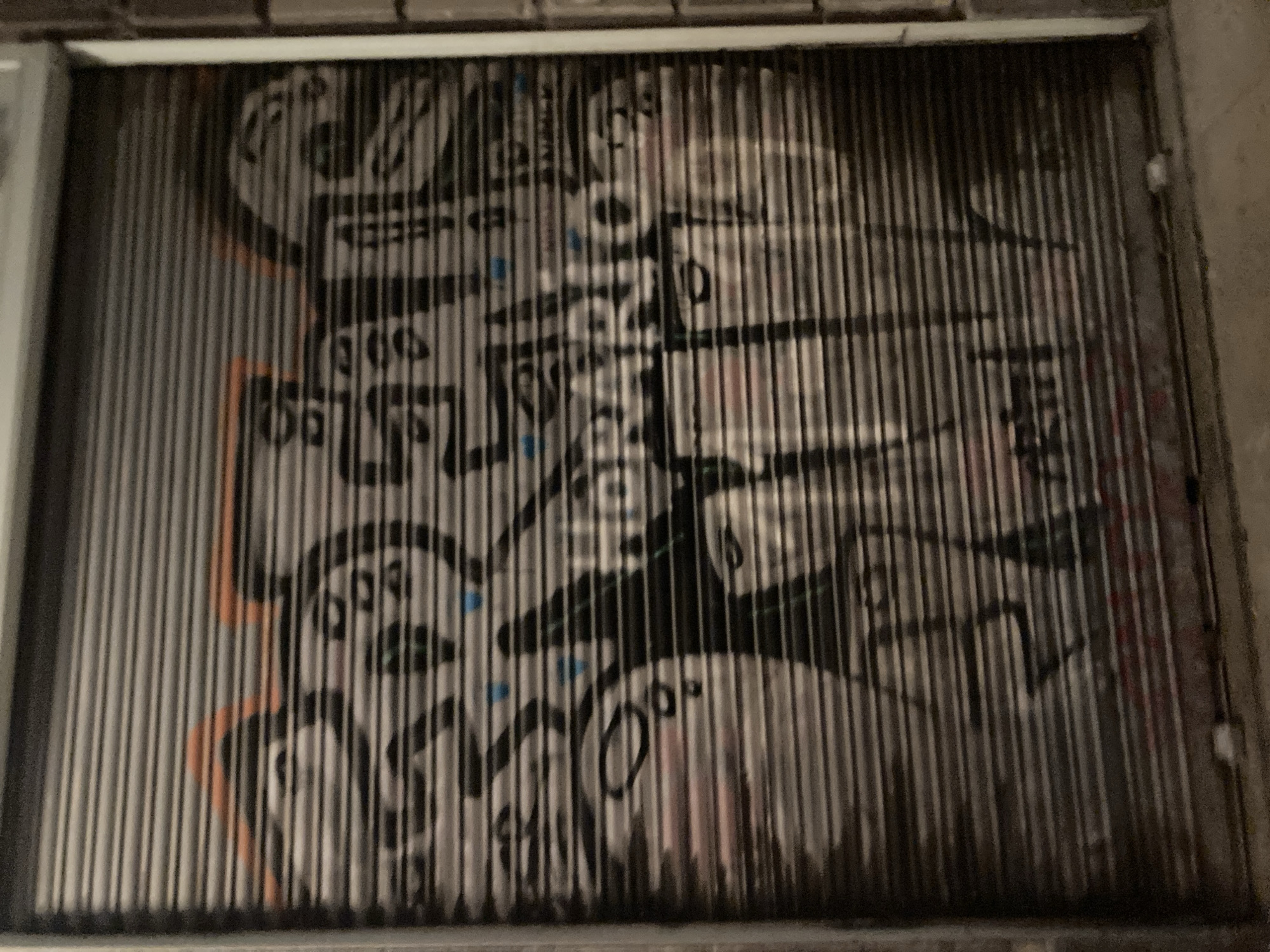 The image shows a graffitied metal roll-up shutter on a building. The graffiti is abstract and features bold lines, figures, and some text. The shutter appears to be dark gray with horizontal slats. Some of the graffiti includes orange accents at the top and blue dots scattered throughout. There are also representations of faces or heads within the graffiti. The scene is dark, suggesting it might be nighttime or an indoor space with artificial lighting. The bottom of the shutter shows some red spray paint and debris on the ground. The building facade to the right of the shutter is light-colored, possibly white or light gray. The overall impression is one of urban street art on a commercial or industrial storefront. The location is identified as Barcelona, Spain.