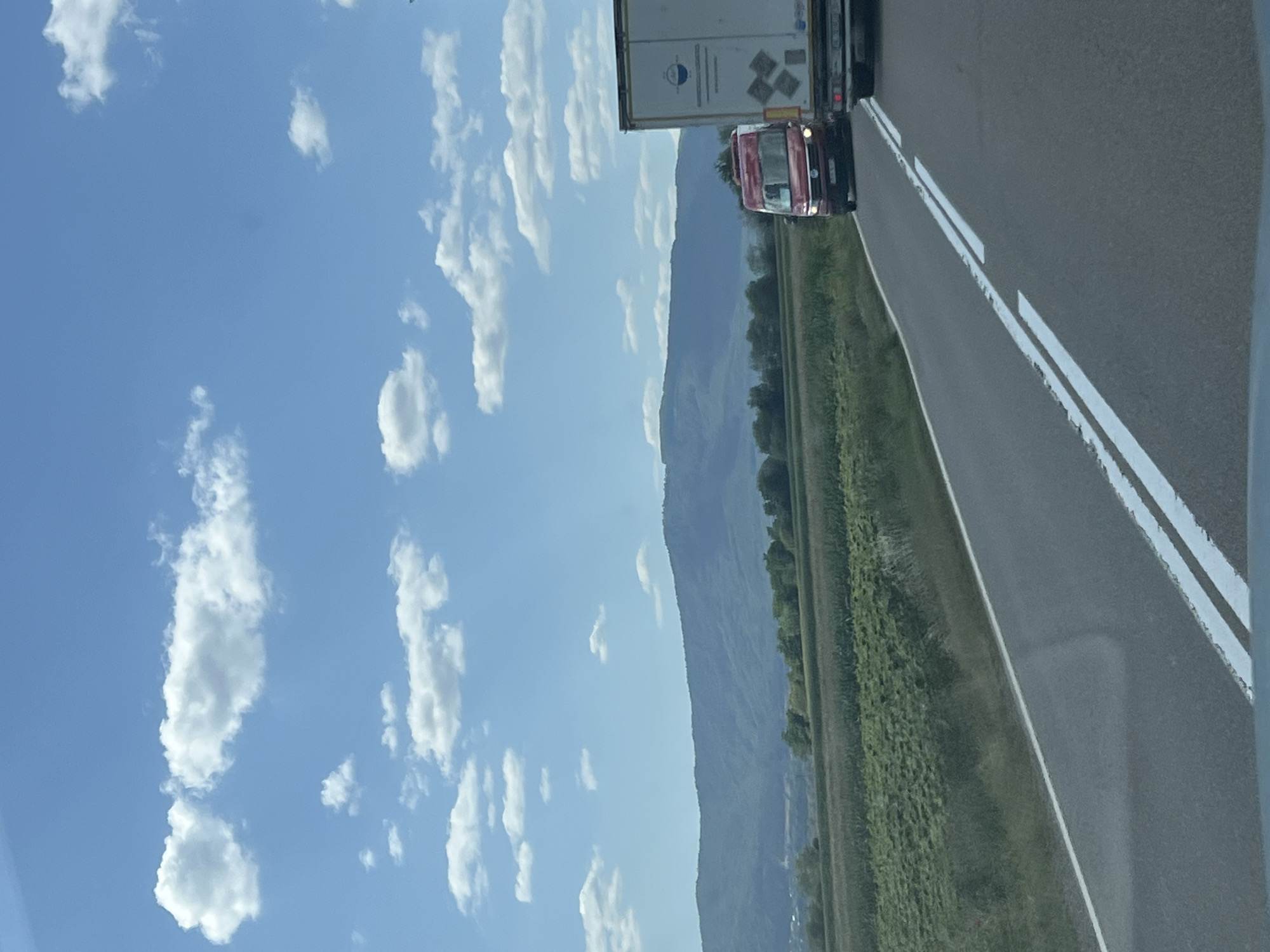 The image shows a two-lane asphalt road with white lane markings, likely in a rural area. On the right side of the road, a white semi-trailer truck is traveling, and to its left, a red van is also on the road. In the background, there are fields of green crops and a range of hills or mountains under a bright blue sky with scattered white clouds. The scene appears to be captured from a vehicle traveling on the road. The location is identified as Berchișești, Romania.