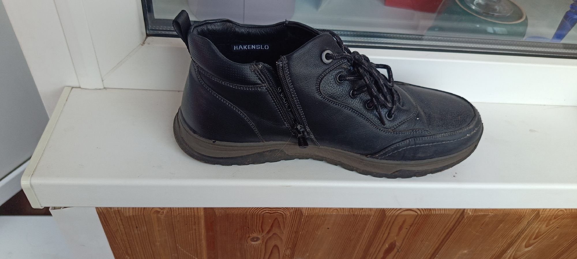 A black leather boot with laces and a side zipper sits on a white windowsill. The boot has the brand name "HAKENSLO" visible inside the collar. The windowsill is positioned above a wooden structure, likely part of a building's exterior. The background shows a window with reflections of what appears to be indoor objects. The scene is indoors, and the specific location within the city of Cheboksary, Russia, cannot be determined from the image alone.