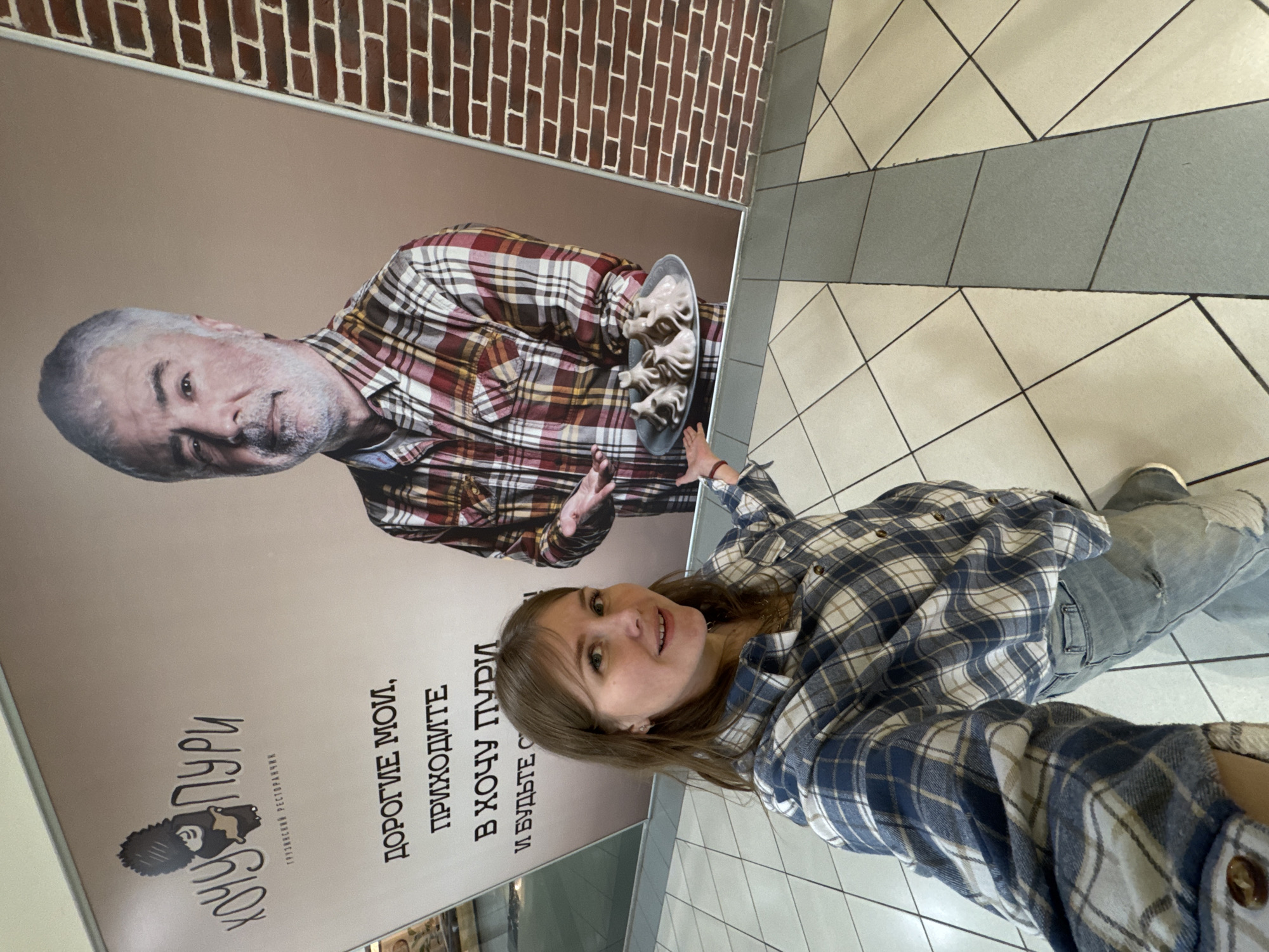 A young woman is captured in a selfie, posing indoors in what appears to be a commercial or public building in Perm, Russia. She stands in front of a large, vertical advertisement for a Georgian restaurant.
The advertisement features a friendly-looking older man with gray hair and a beard, wearing a red, white, and brown plaid shirt. He is holding a plate piled with what appear to be Georgian khinkali (dumplings), extending them towards the viewer. A brick pattern is visible in the upper part of the ad, above the man's head, while the rest of the background is a solid light brown.
The woman in the foreground wears a blue, white, and tan plaid shirt, mirroring the man's attire in the ad, and light-colored jeans. She looks at the camera with a pleasant, smiling expression. Her right arm is outstretched, and she points directly at the food held by the man in the advertisement, creating a playful interaction with the promotional material.
The floor is made of light and dark gray tiles arranged in a diagonal pattern. The lighting suggests an indoor daytime setting.
The visible text on the advertisement is in Russian. The restaurant's logo reads "ХОЧУ ПУРИ" (Khochu Puri), translating to "I Want Puri" (Puri being a type of Georgian bread). Below the logo, it's explicitly identified as a "ГРУЗИНСКИЙ РЕСТОРАНЧИК" (Georgian Restaurant). A larger inscription next to the man invites patrons with "ДОРОГИЕ МОИ, ПРИХОДИТЕ В ХОЧУ ПУРИ И БУДЬТЕ С..." (My dears, come to Khochu Puri and be...), with the final word cut off. The scene portrays a casual, engaging moment with an advertisement for Georgian cuisine.